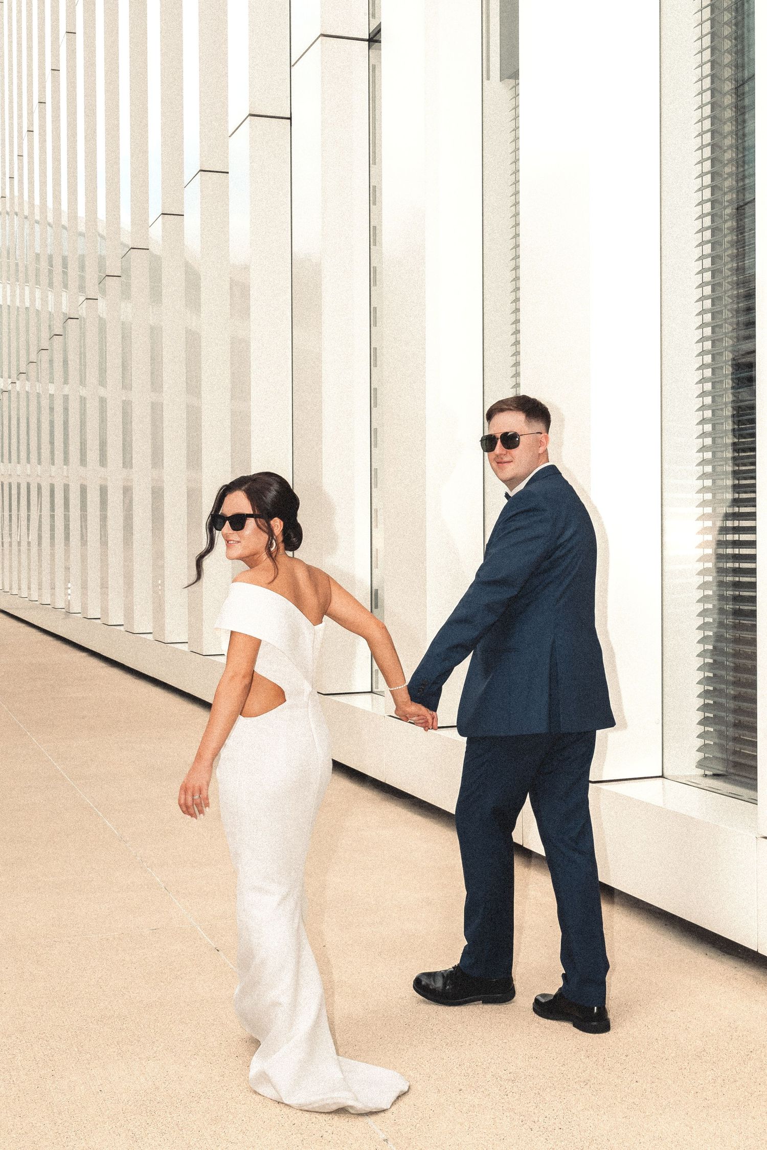 Bride and groom walking hand in hand in a sun-drenched colonnade, sharing a joyful moment.