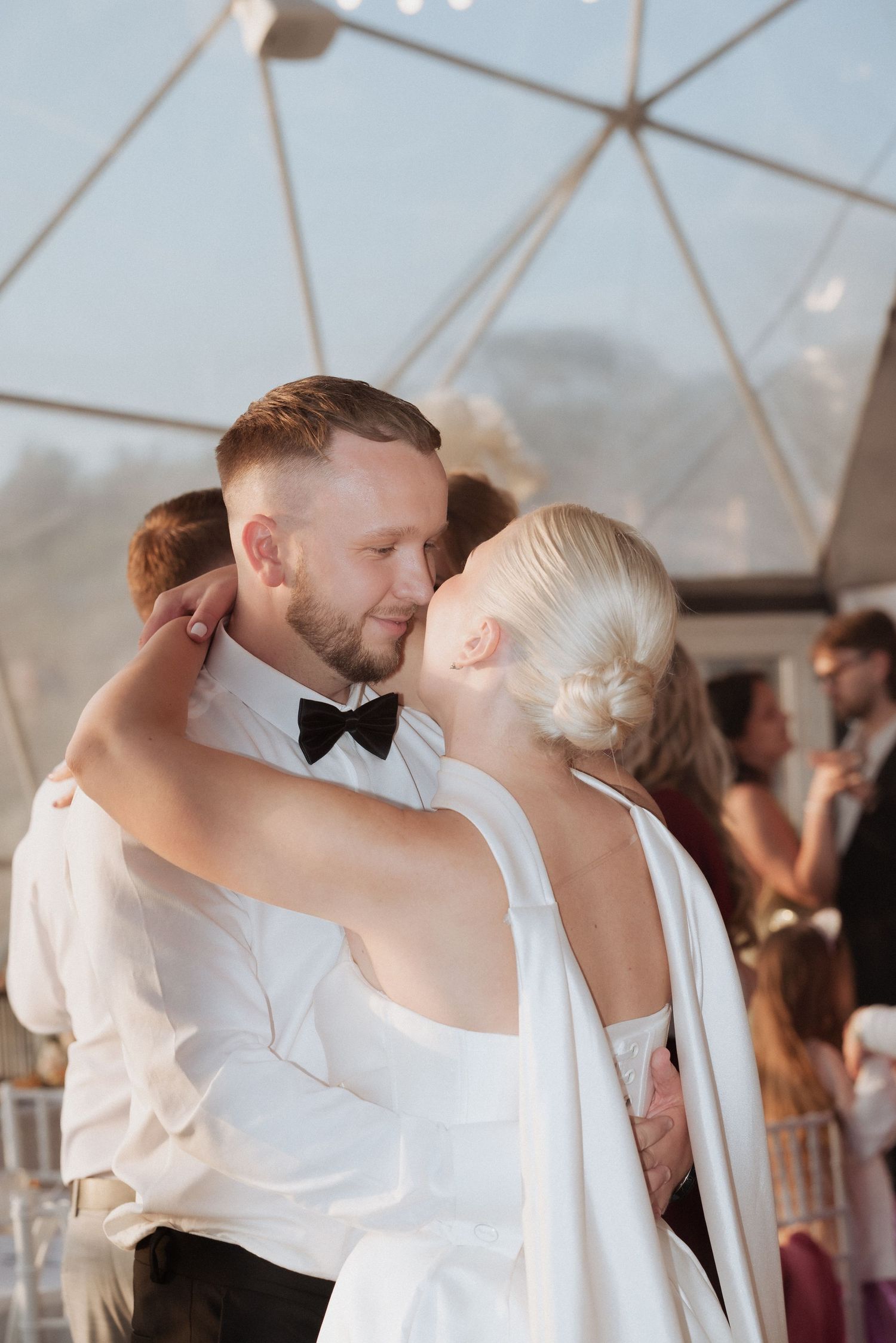 Bride and groom sharing a kiss under a tented wedding reception in Klaipėda, Lithuania, surrounded by joyful guests.