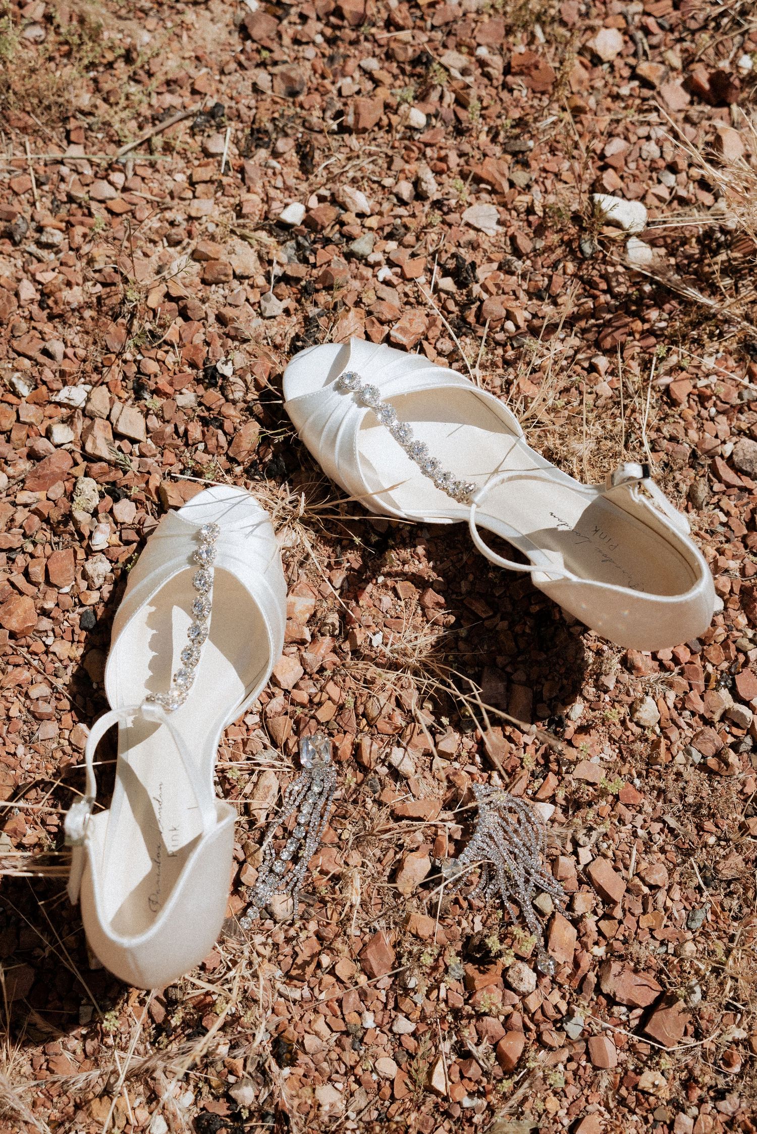 White bridal shoes with bows resting on red gravel, styled for a flat lay detail shot in natural light.