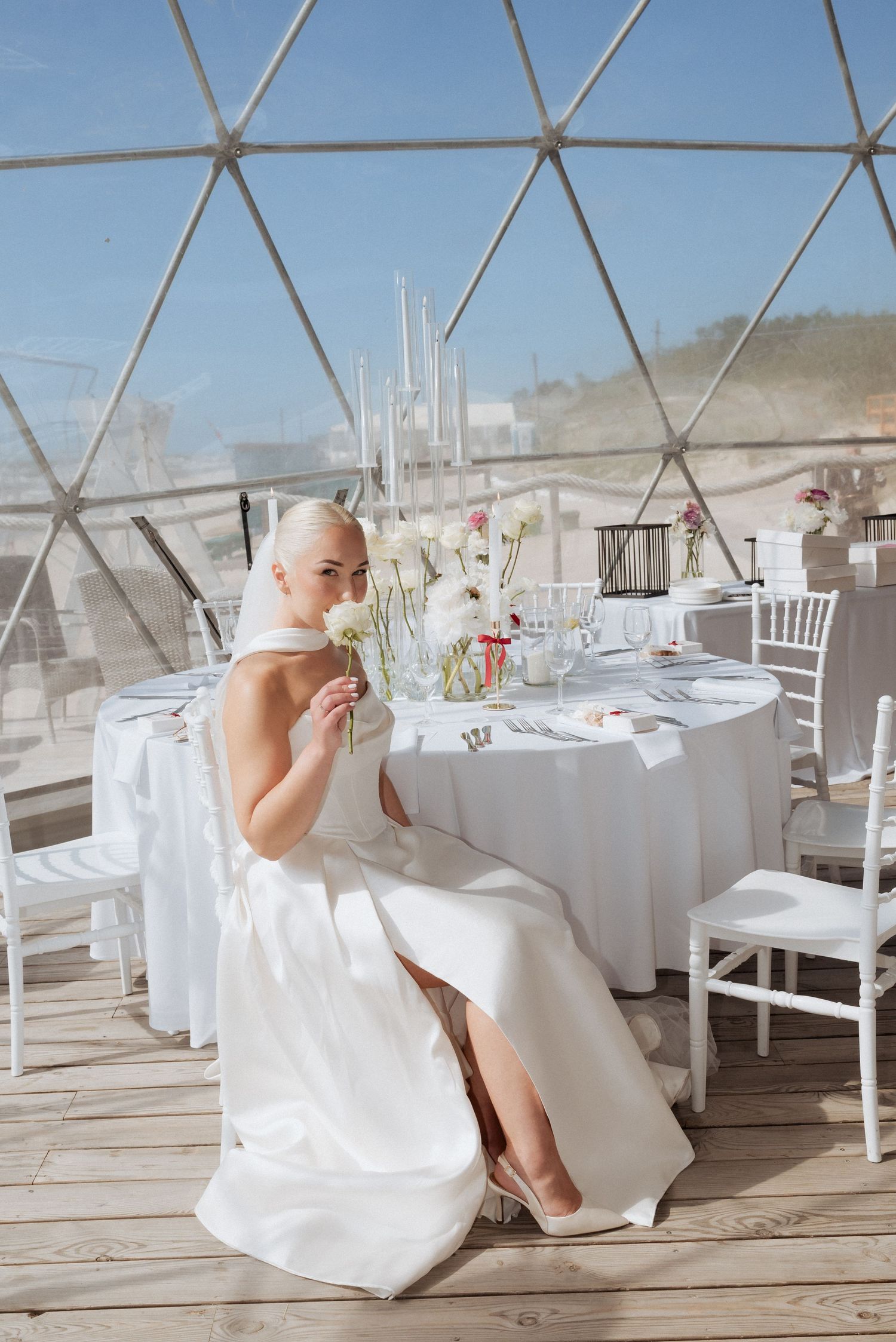 Bride seated at a reception table under a transparent dome setup, surrounded by elegant decor and natural light.