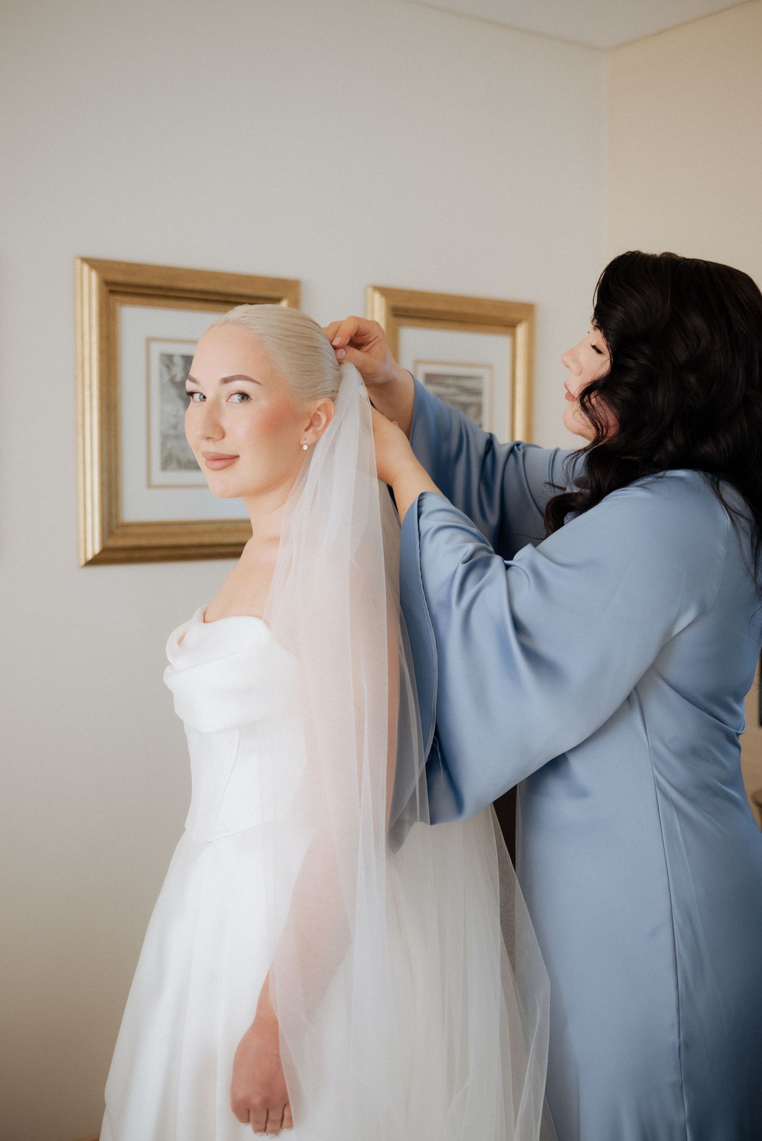 Bride mother helping the bride with final dress adjustments in a warmly lit room before the ceremony.