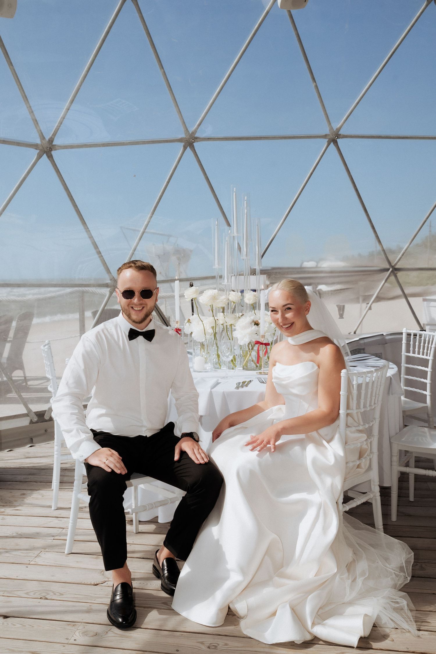 Bride and groom sitting side by side under a modern tent structure during their stylish outdoor wedding celebration.