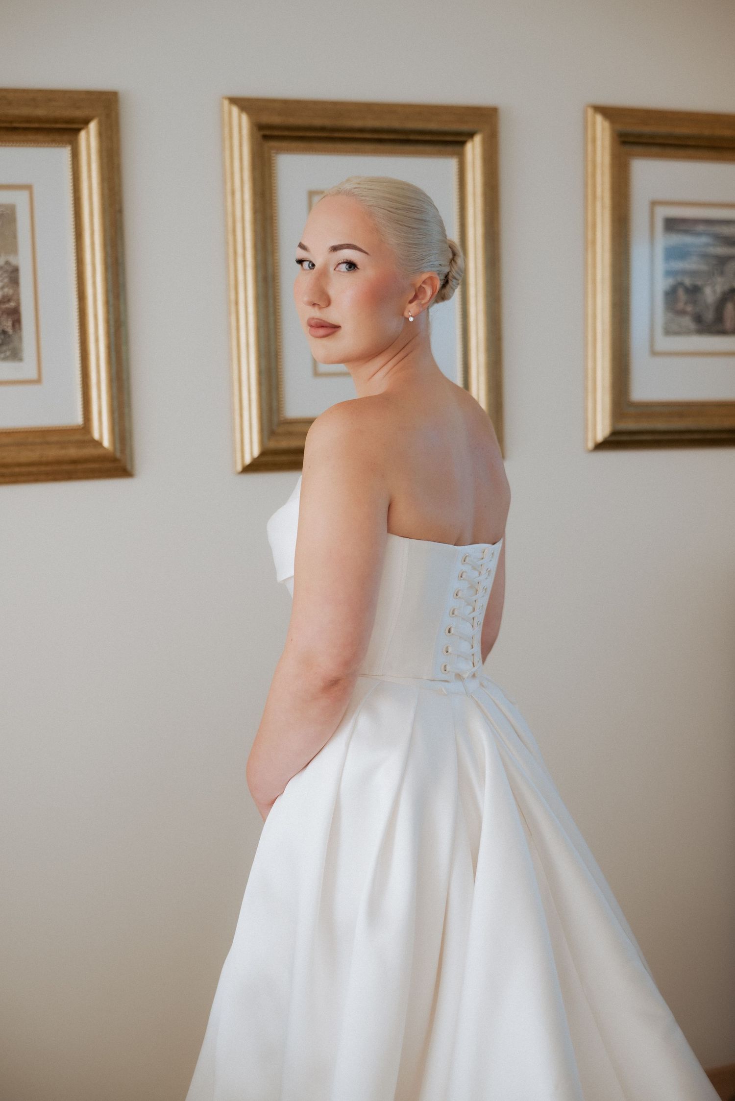 Bride adjusting her dress while standing in front of a framed painting, bathed in soft natural light.