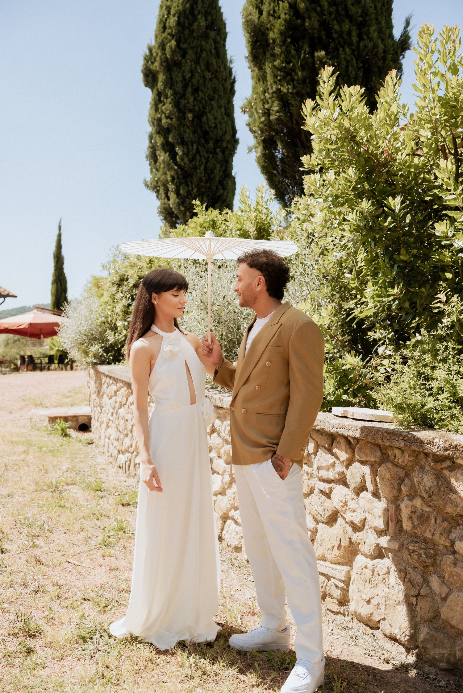 Bride and groom walking toward the camera through a Tuscan garden, surrounded by olive trees and stone walls with paper parasol.