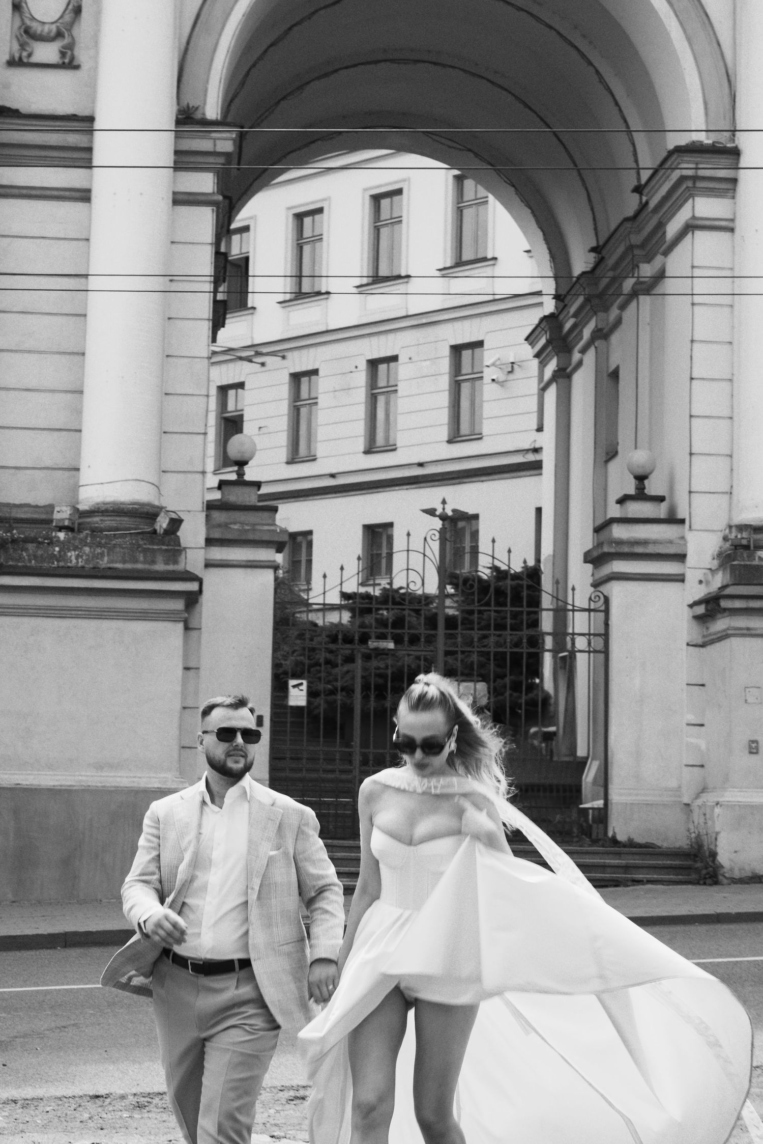 juodai balta fotografija, fone didelė arka, o nuotaka ir jauniki susikibę eina gatve