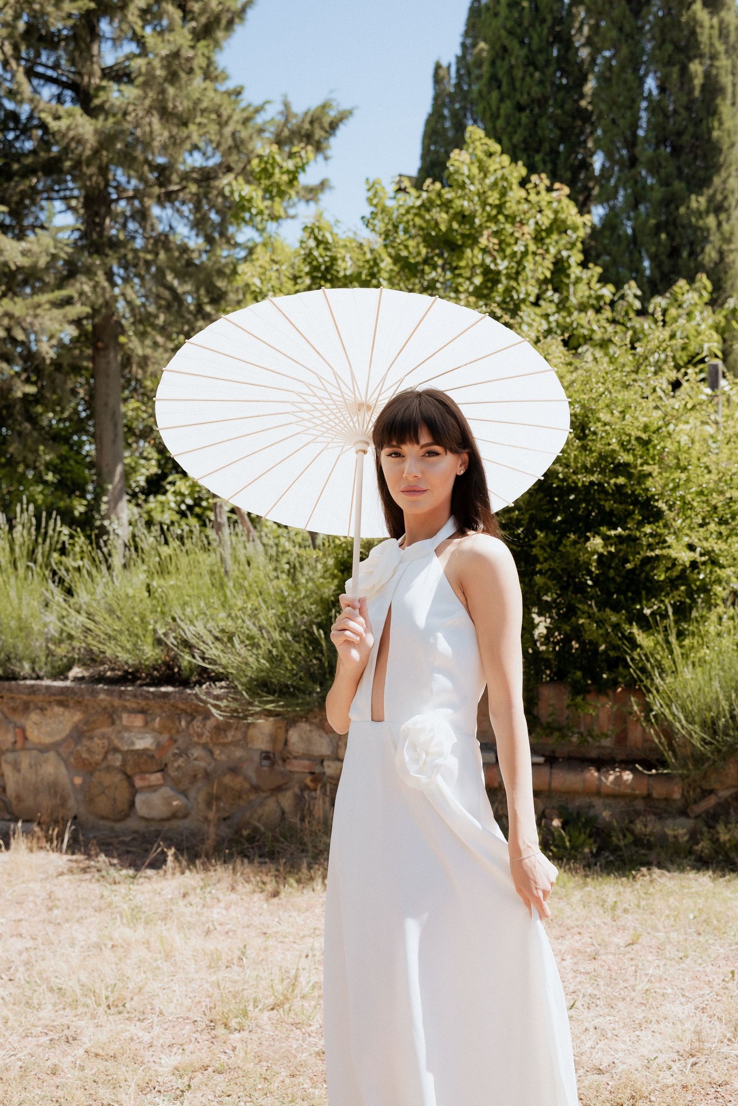 Bride standing alone with a white parasol in hand, set against a rustic Tuscan landscape.