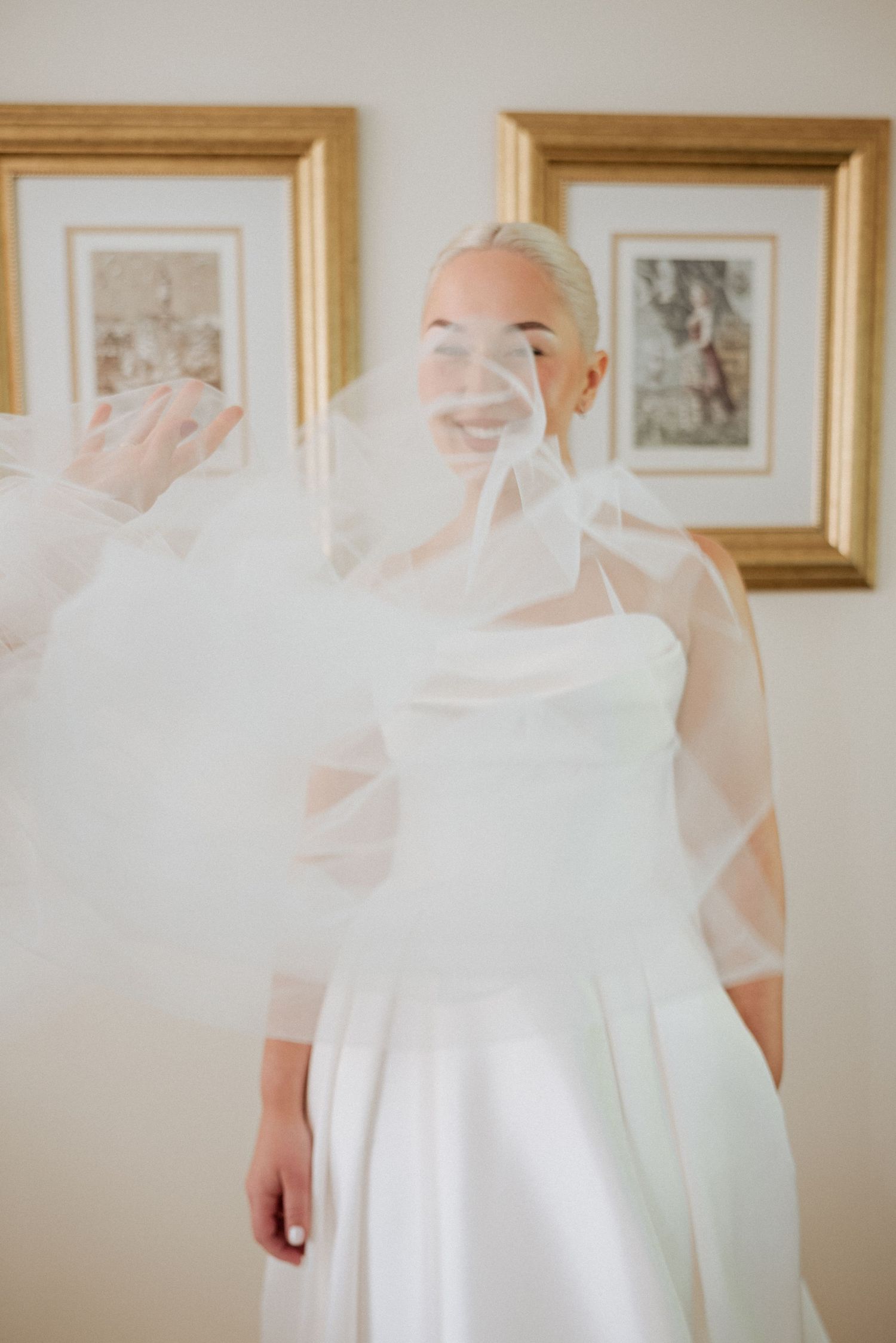 Bride with her veil softly floating in front of a framed artwork, captured in a peaceful indoor bridal portrait.