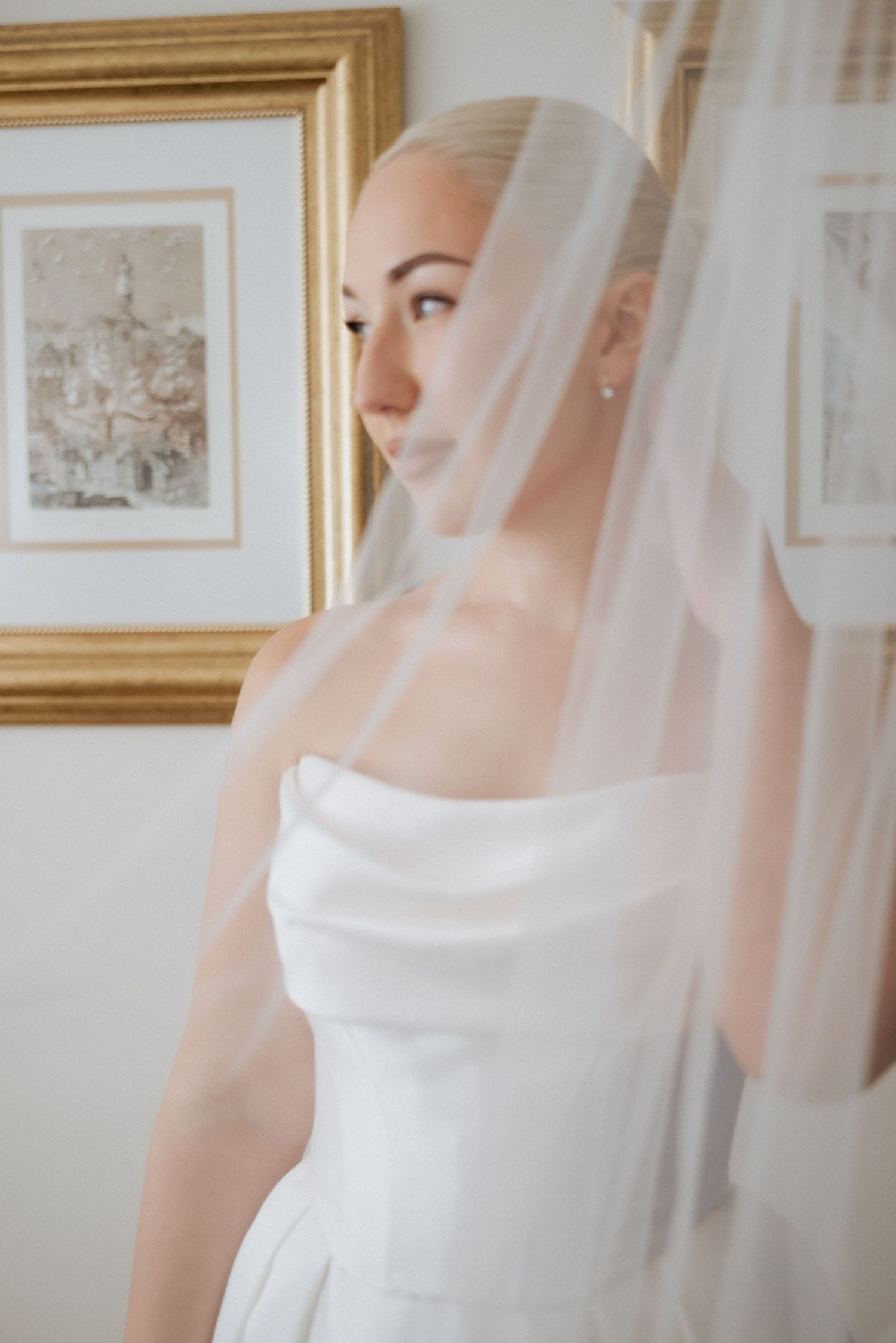 Bride captured through a soft veil in a quiet, artistic bridal portrait near a framed artwork.