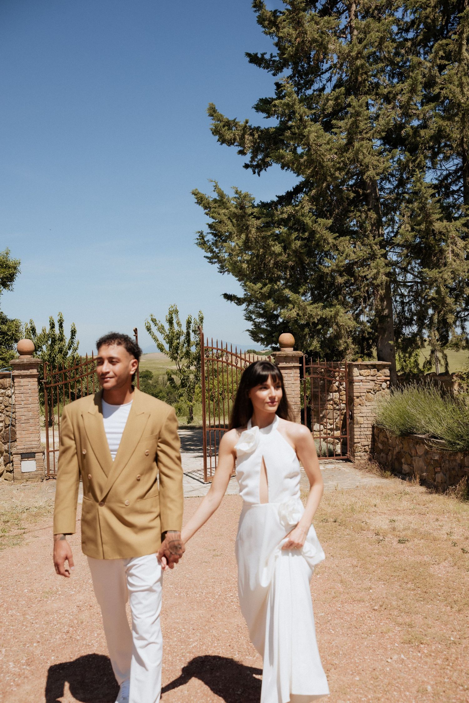 Bride and groom walking through a Tuscan garden pathway, surrounded by olive trees and rustic charm.