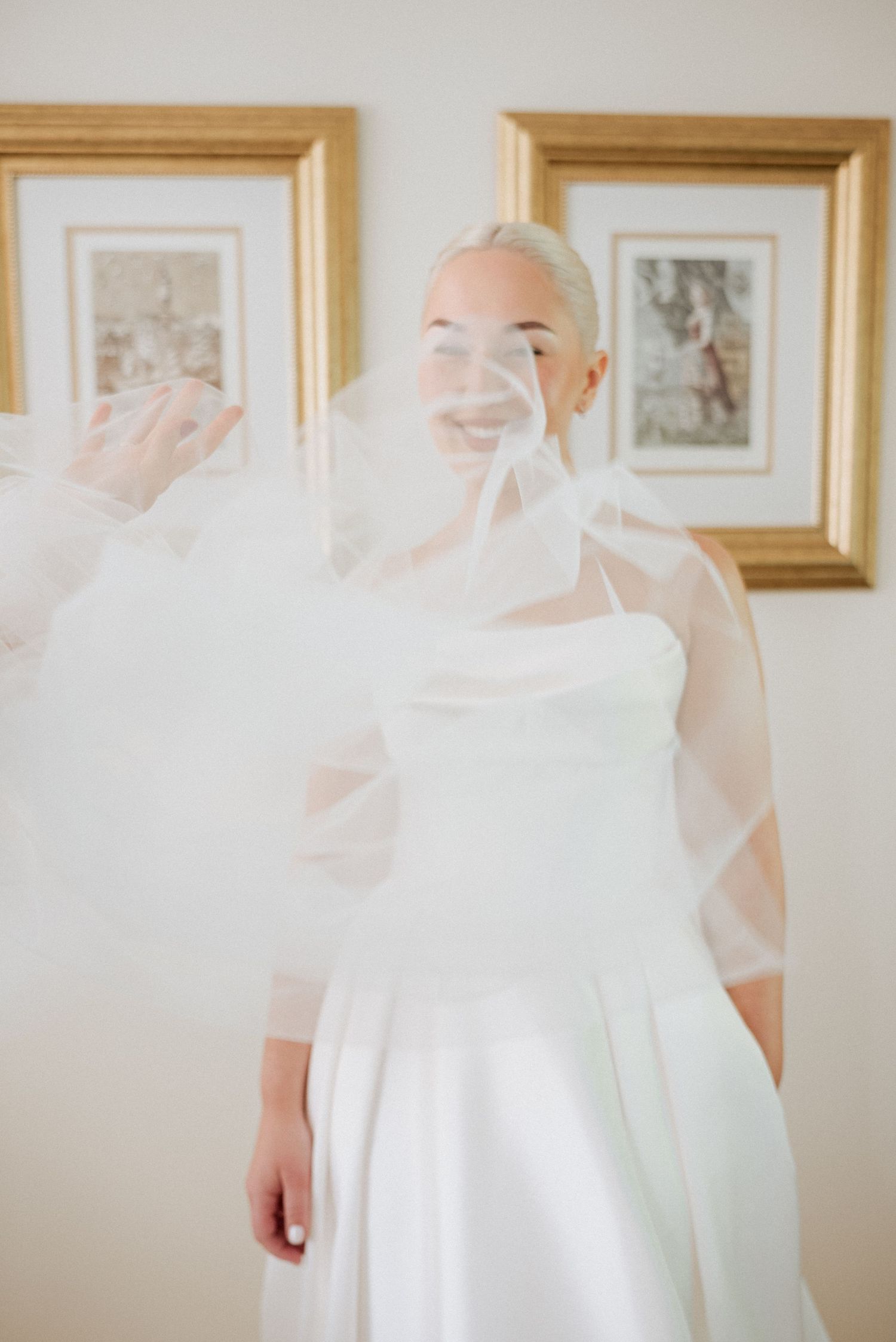 Bride standing with veil billowing in front of framed art, captured in a calm and elegant pre-ceremony moment.