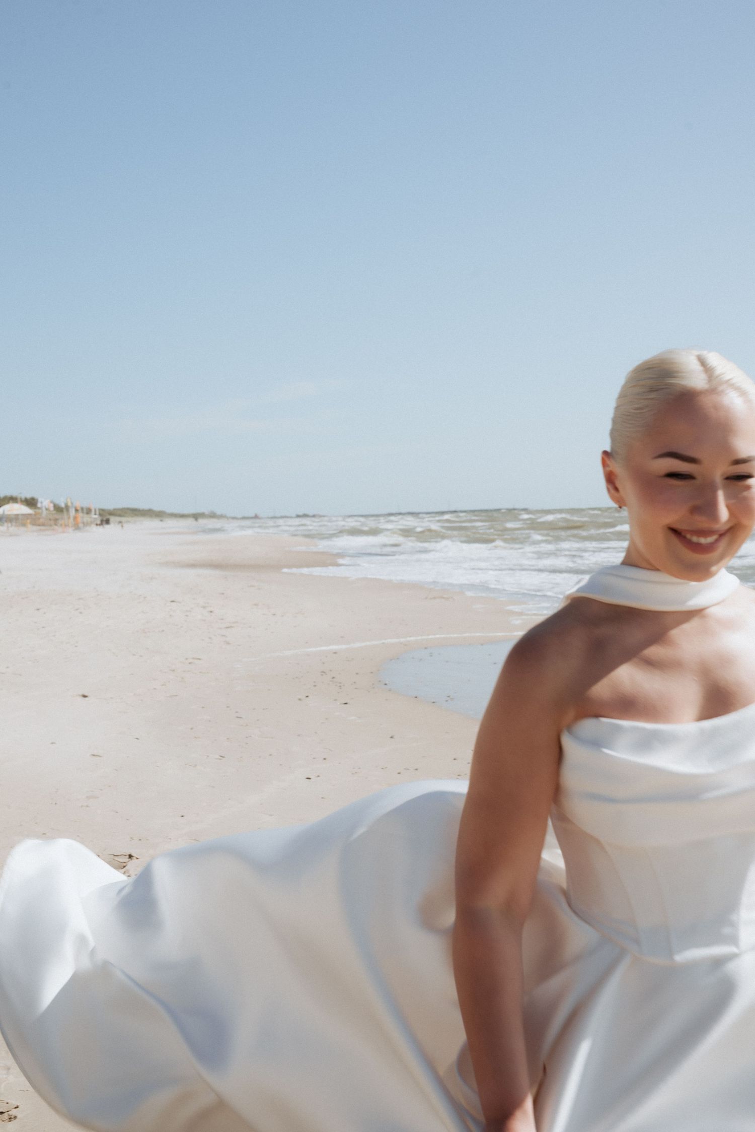 Bride sitting on the sand in her wedding dress, smiling softly with the sea view stretching behind her.
