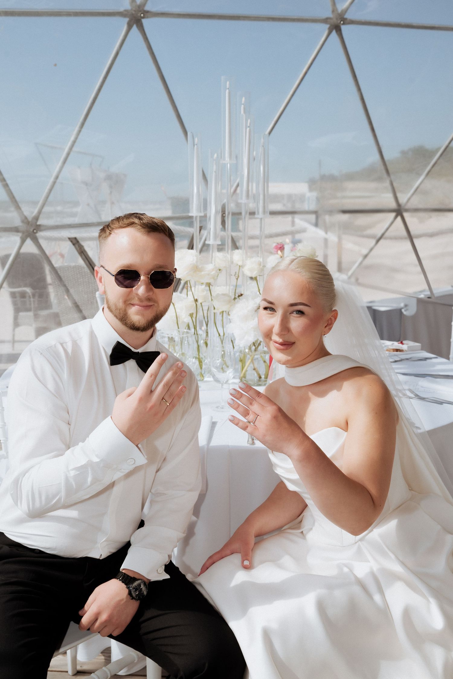 Groom seated and bride laughing beside him under a transparent tent structure during an outdoor wedding reception.