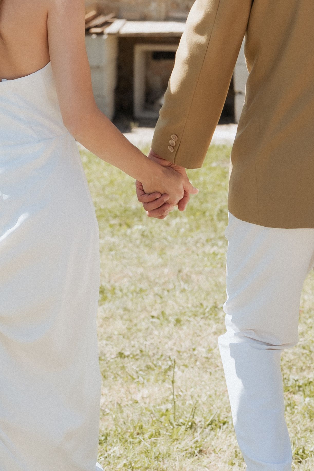 Bride and groom holding hands, walking away from the camera across a sunlit field in Tuscany.