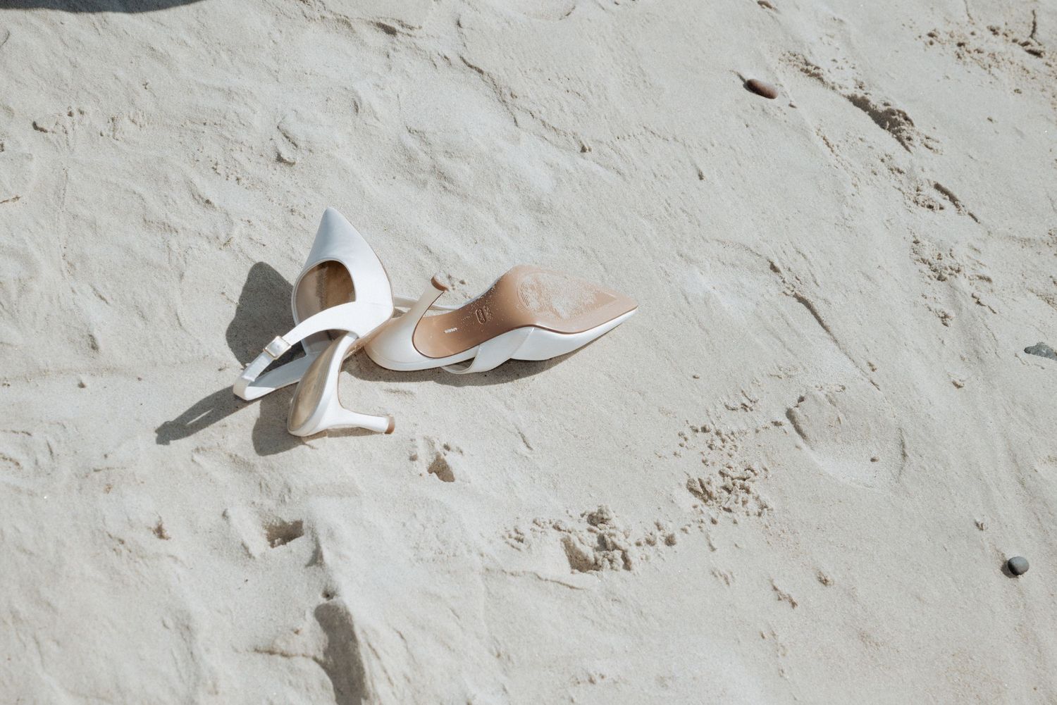 A pair of bridal shoes left in the sand, photographed on a quiet beach with sunlight and soft shadows.