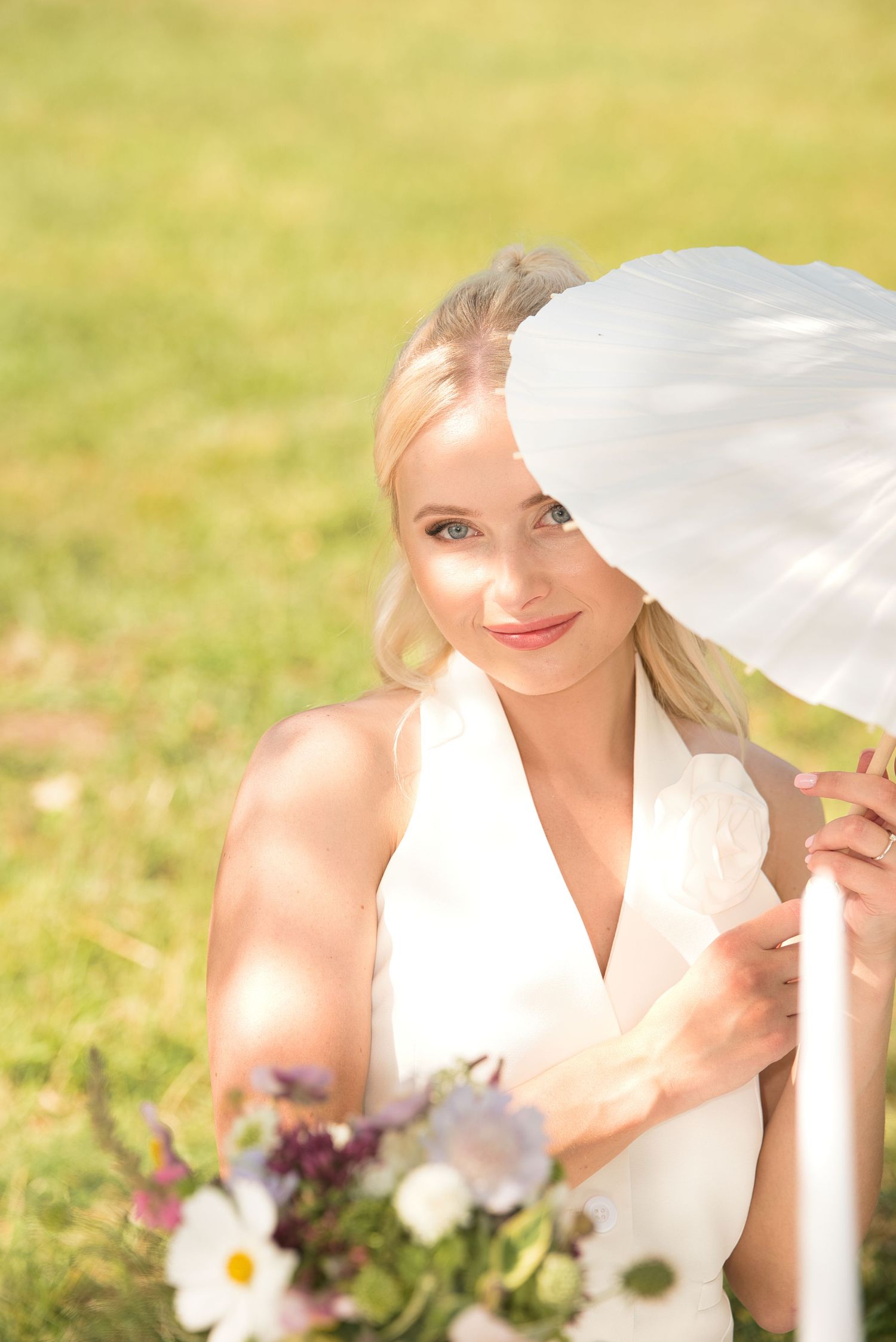 Bride sitting outdoors in natural light, partially shaded by parasol and flowers nearby.
