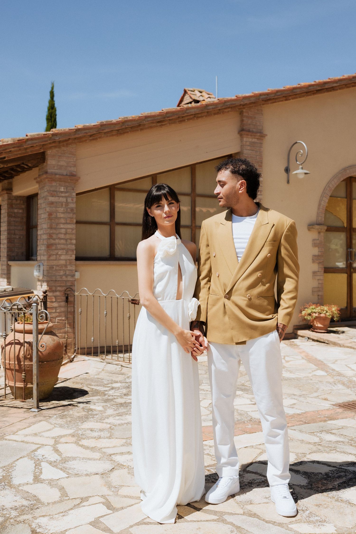 Bride and groom standing outside a terracotta-colored Tuscan villa, posing for a portrait in full sun.