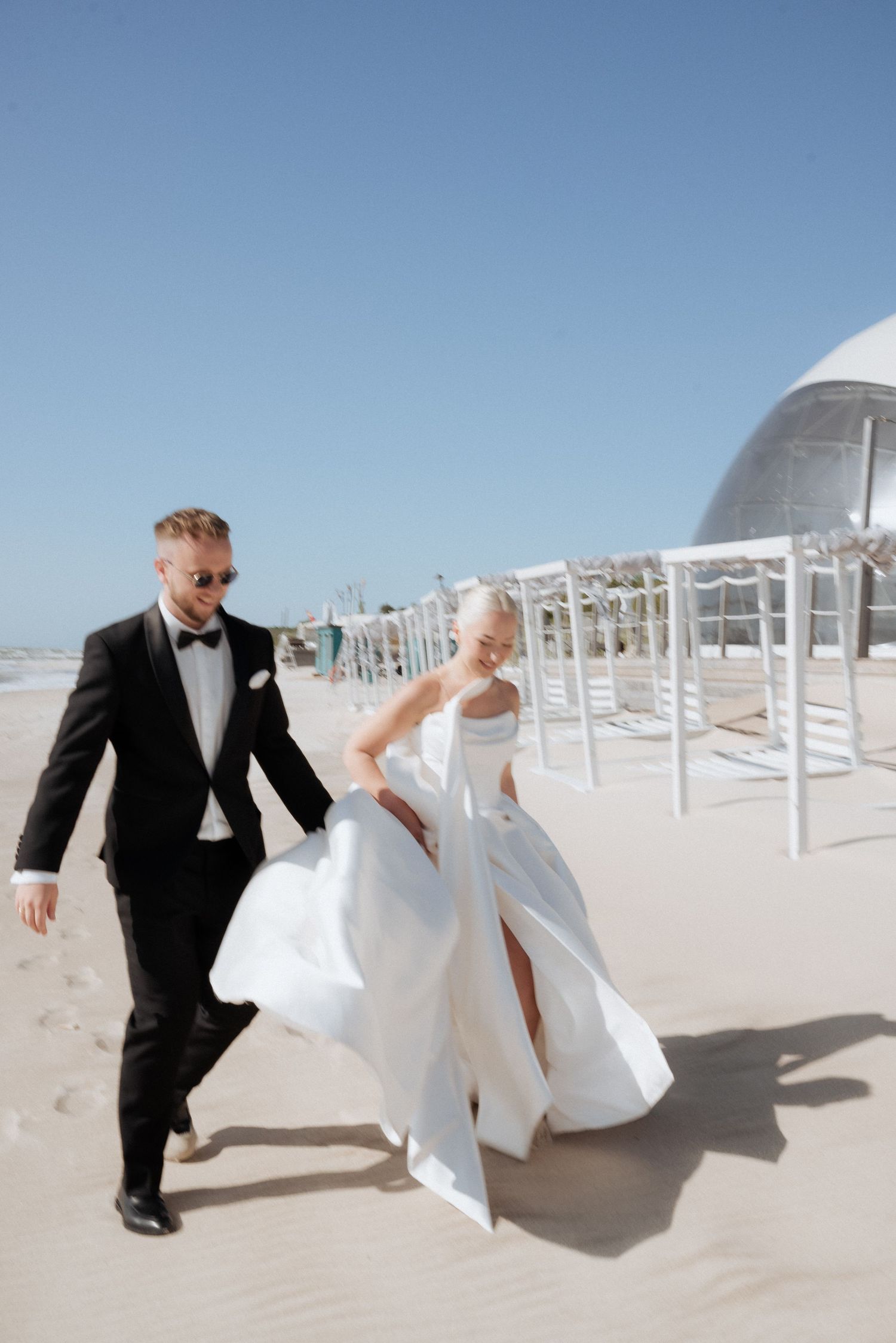 Bride and groom walking hand in hand along the shore with the sea in the background, captured on a bright beach day.