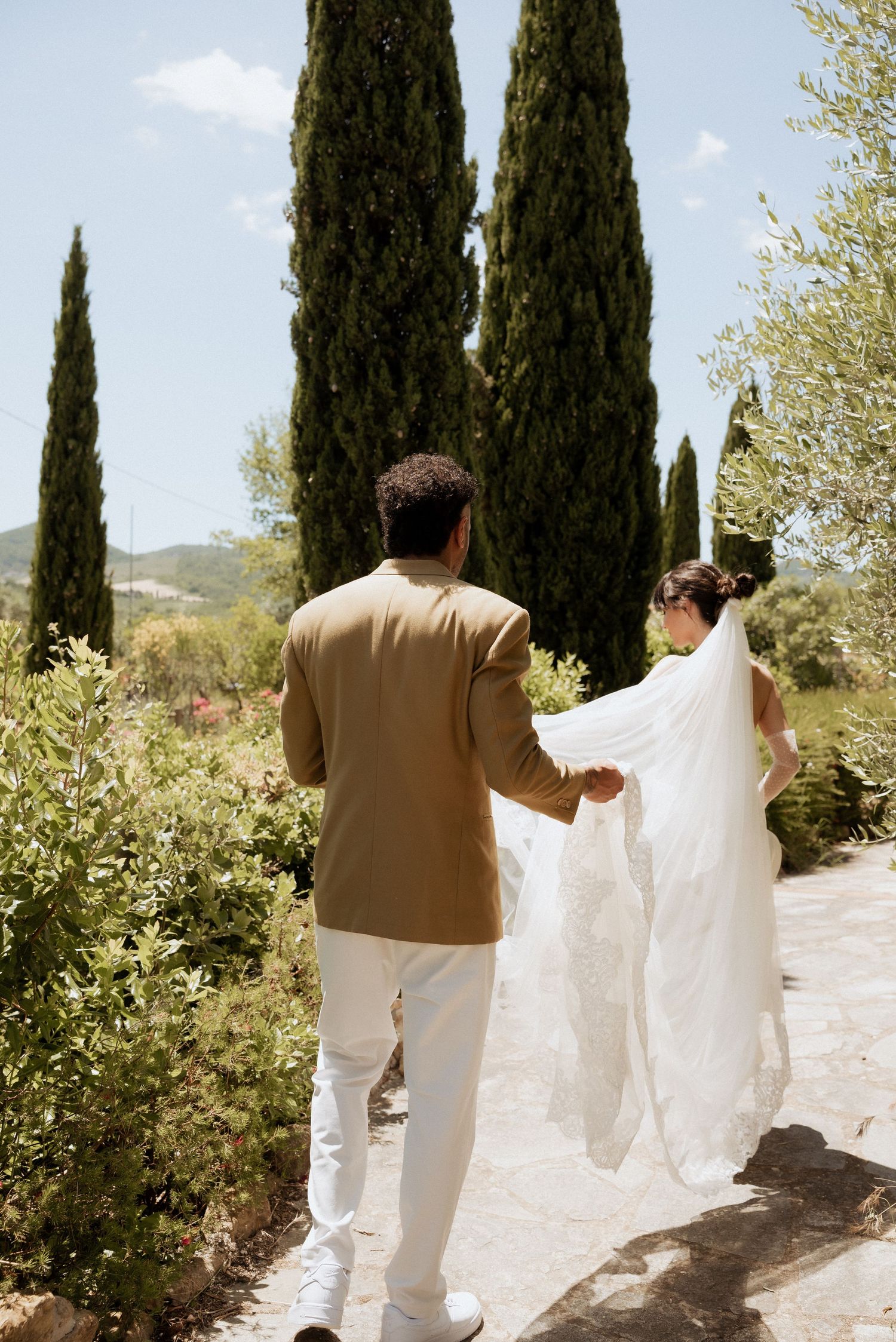 Bride and groom walking side by side down a sunlit cypress-lined path in the Tuscan countryside.