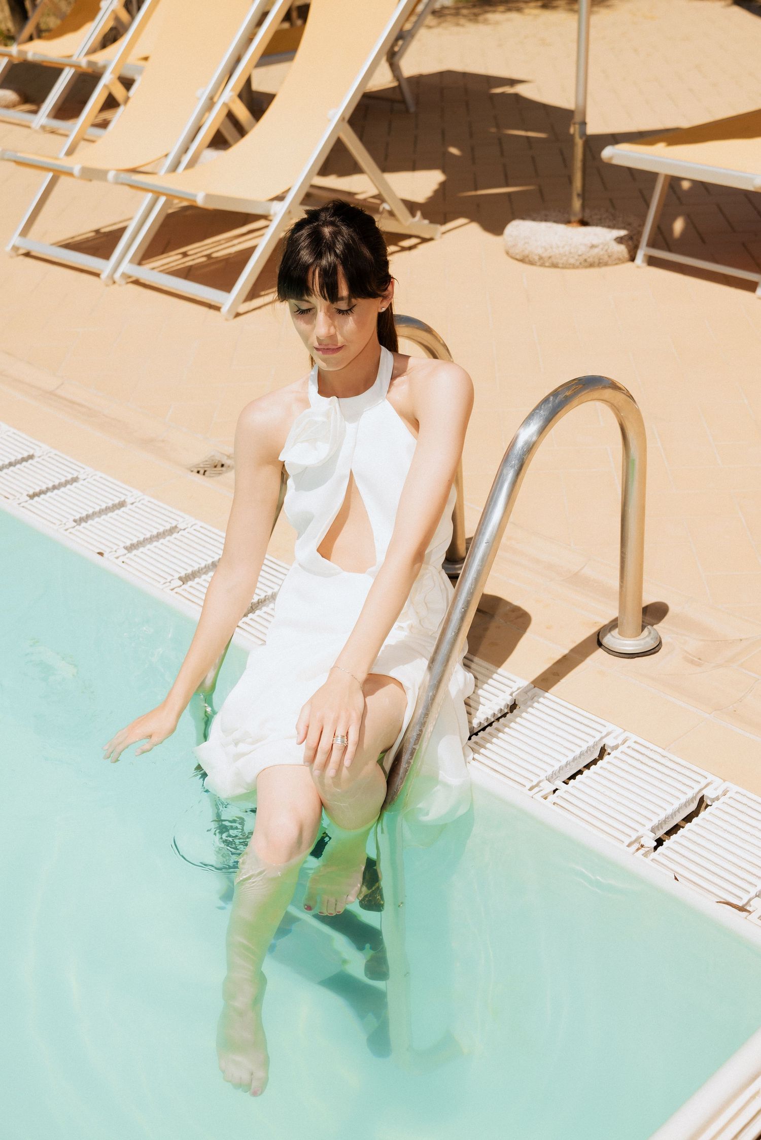 Bride sitting at the edge of a turquoise pool in a white dress, dipping her feet into the water on a sunny day.