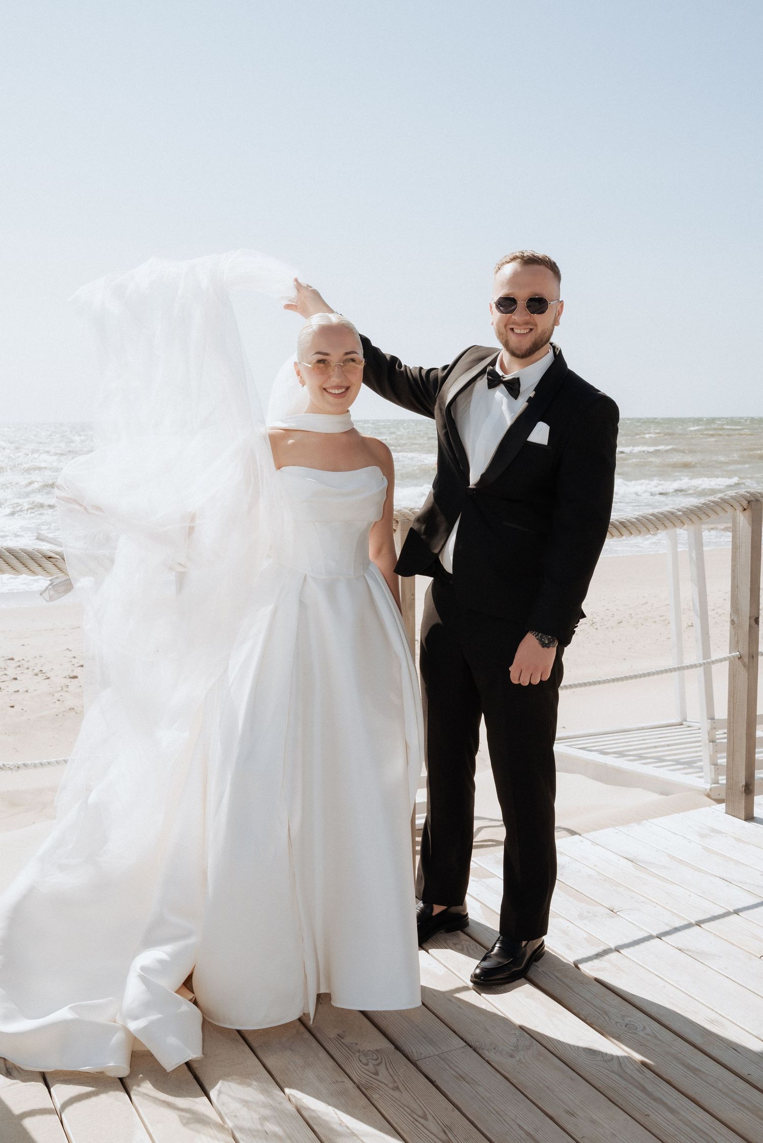 Bride and groom holding hands and smiling as they walk along a coastal boardwalk with the sea behind them.