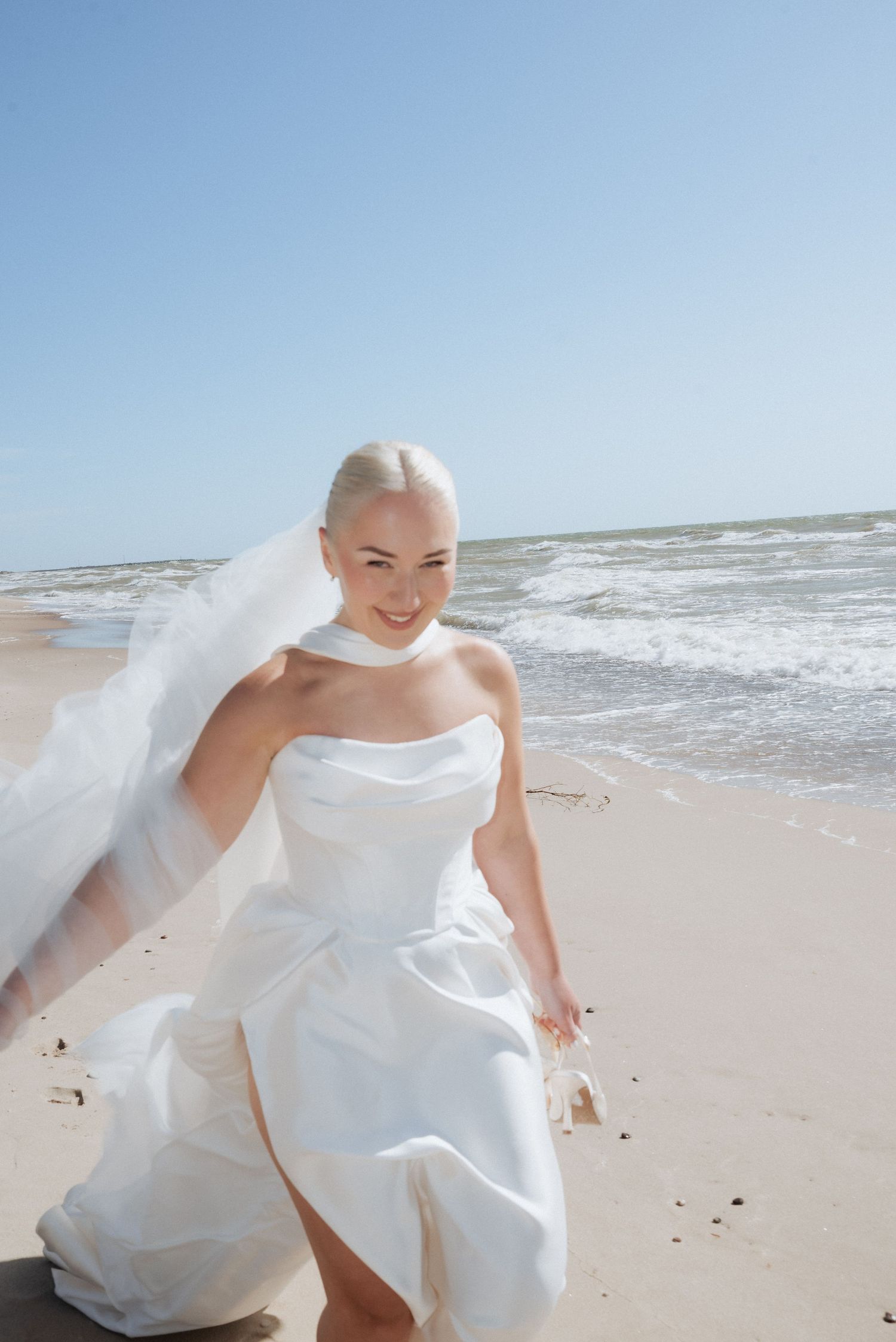 Bride walking barefoot along the shoreline in her gown, the ocean sparkling behind her in a relaxed beach wedding moment.
