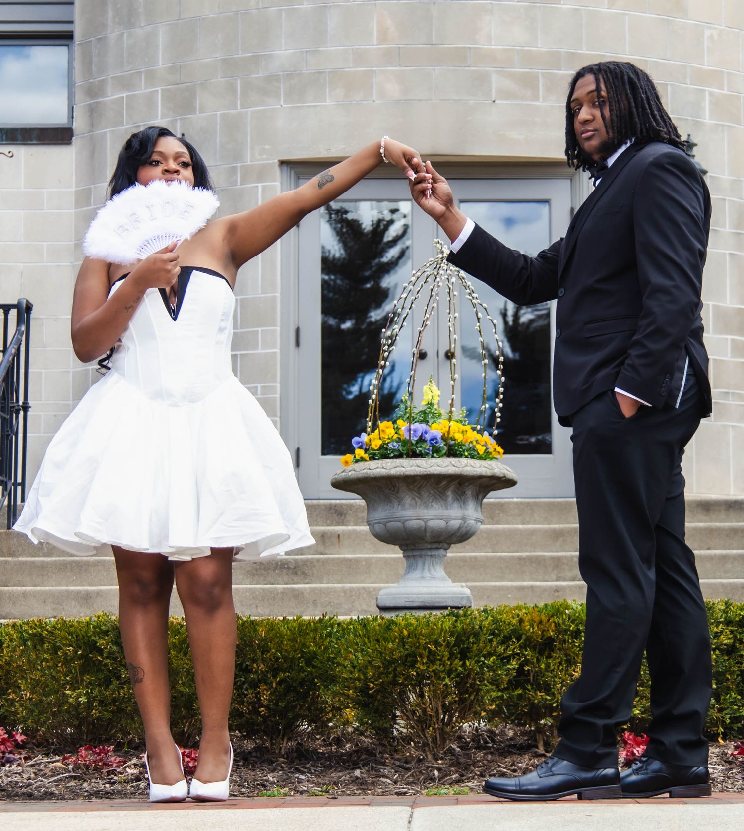 A couple in formal attire poses playfully with a white fan near a decorative garden urn.