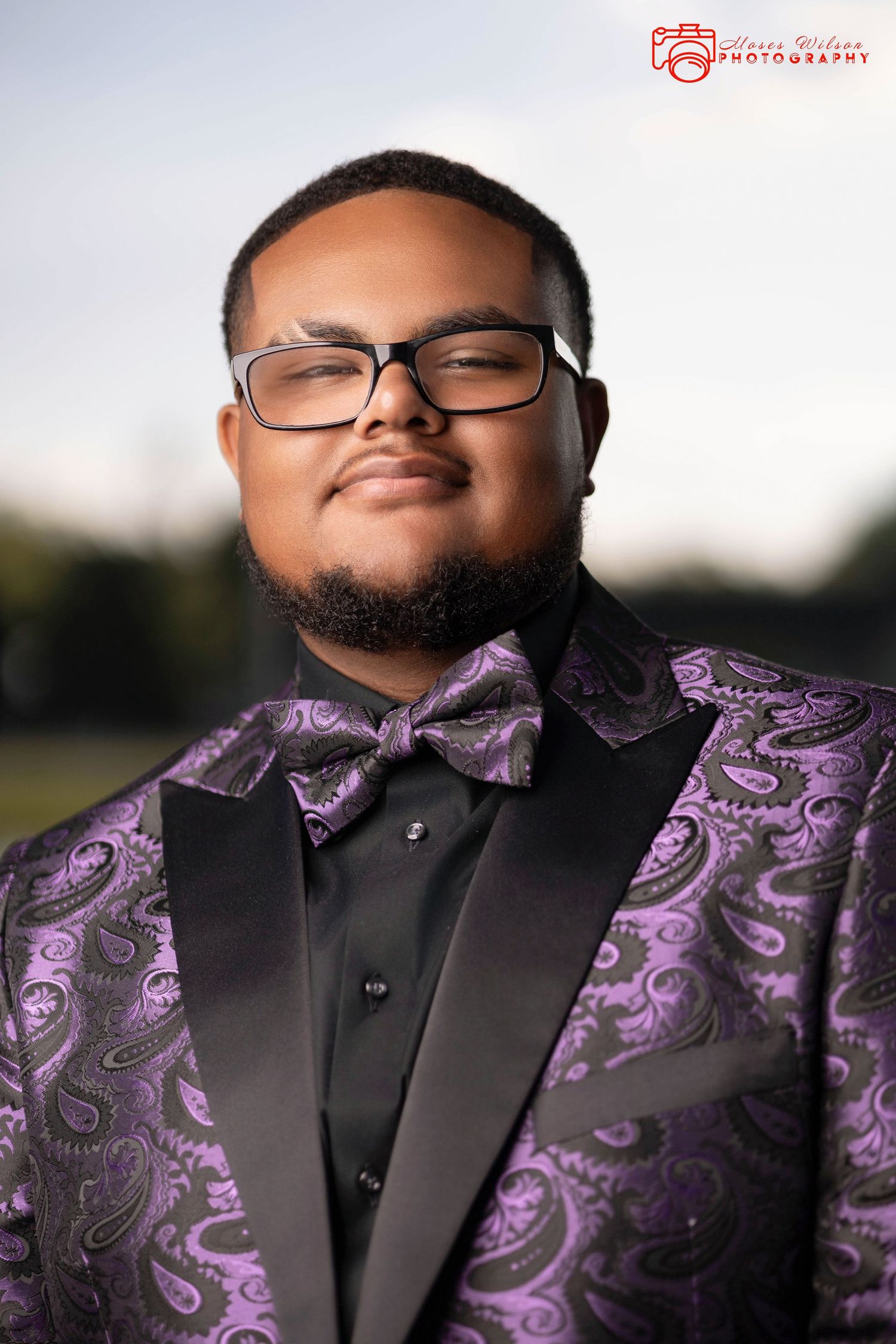 Young man in purple paisley tuxedo jacket with black bow tie and glasses poses outdoors for formal portrait.