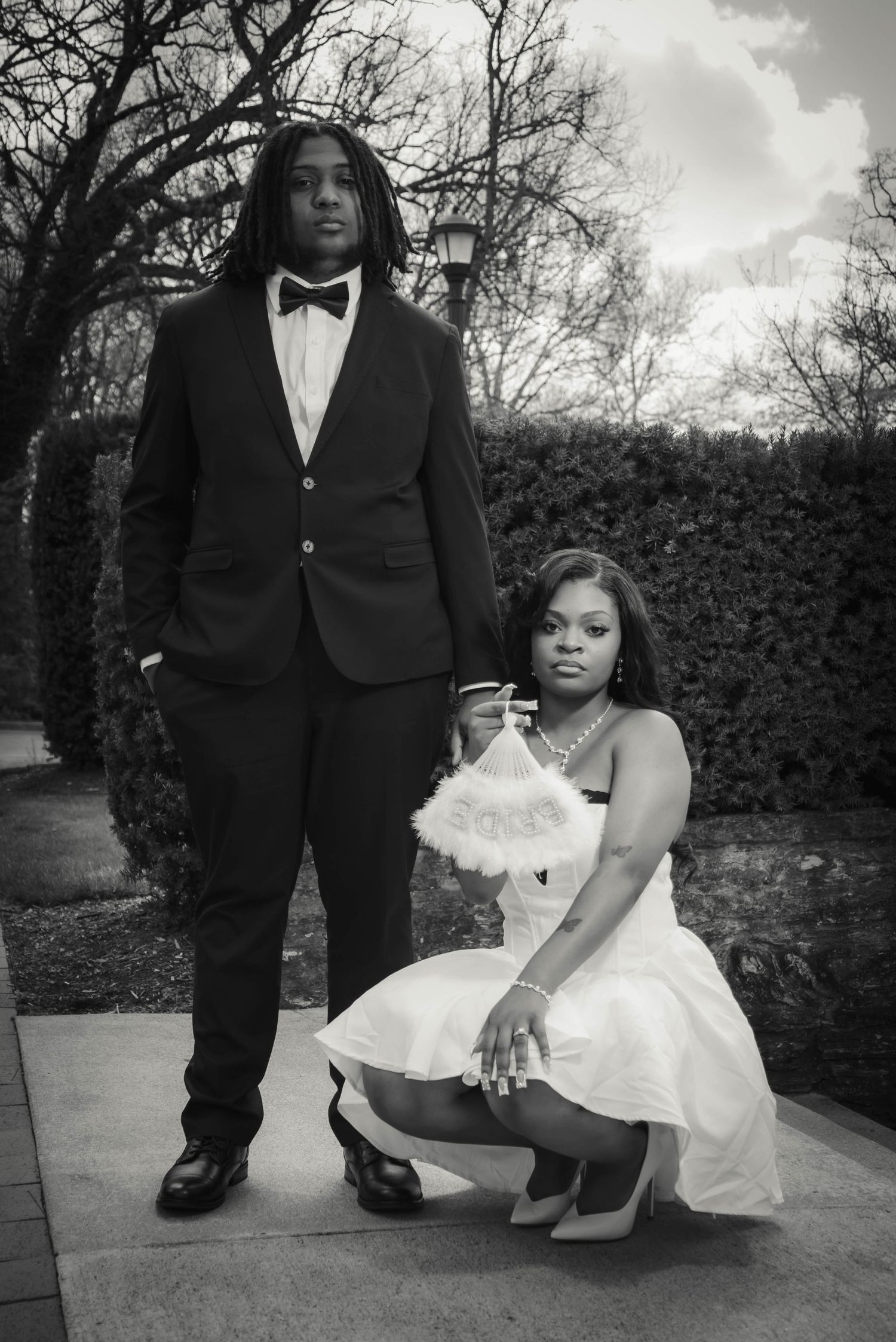 A stylish couple poses for a black and white portrait near hedges.