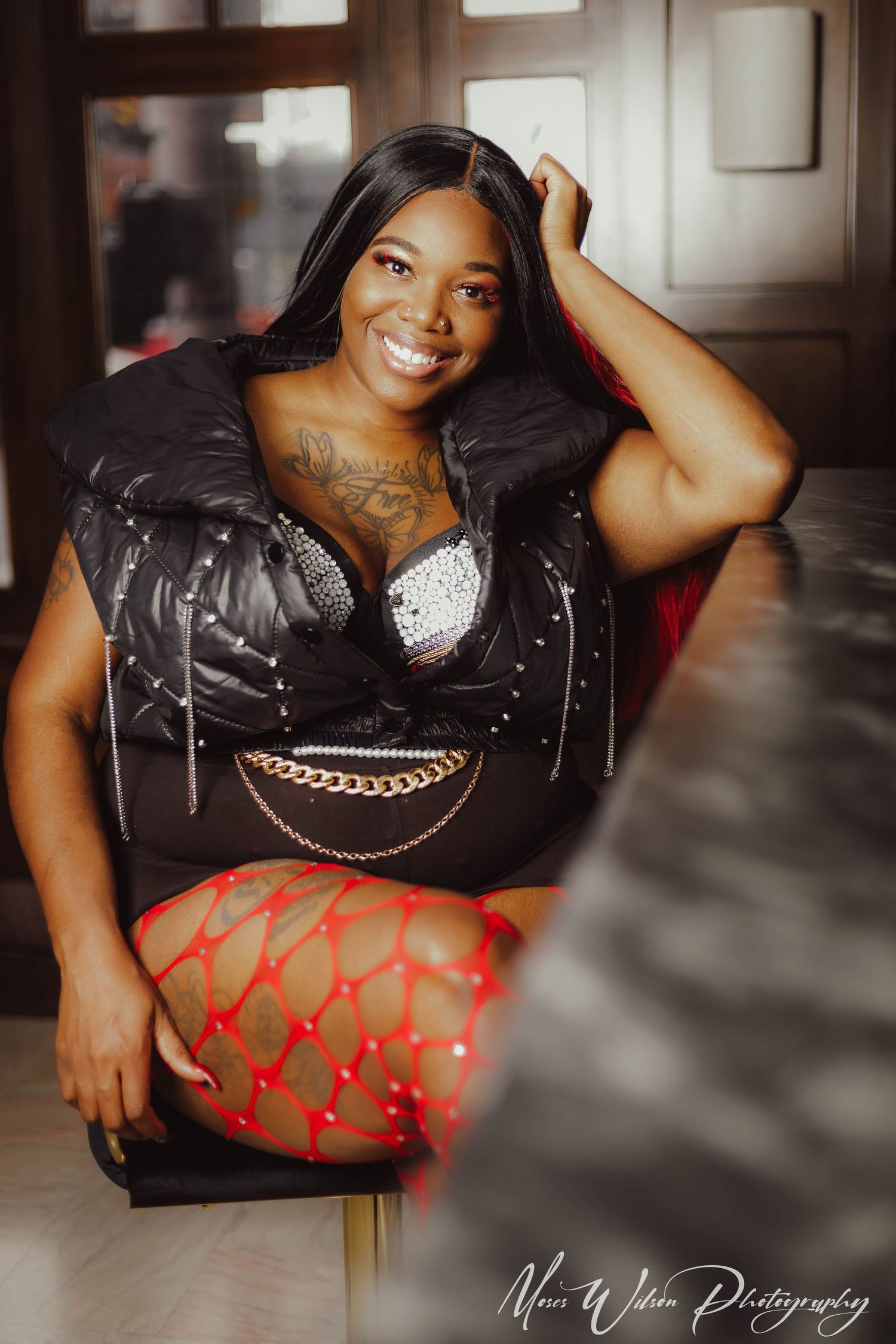 Professional boudoir portrait in a black leather top and red patterned fishnet stockings against moody indoor lighting.