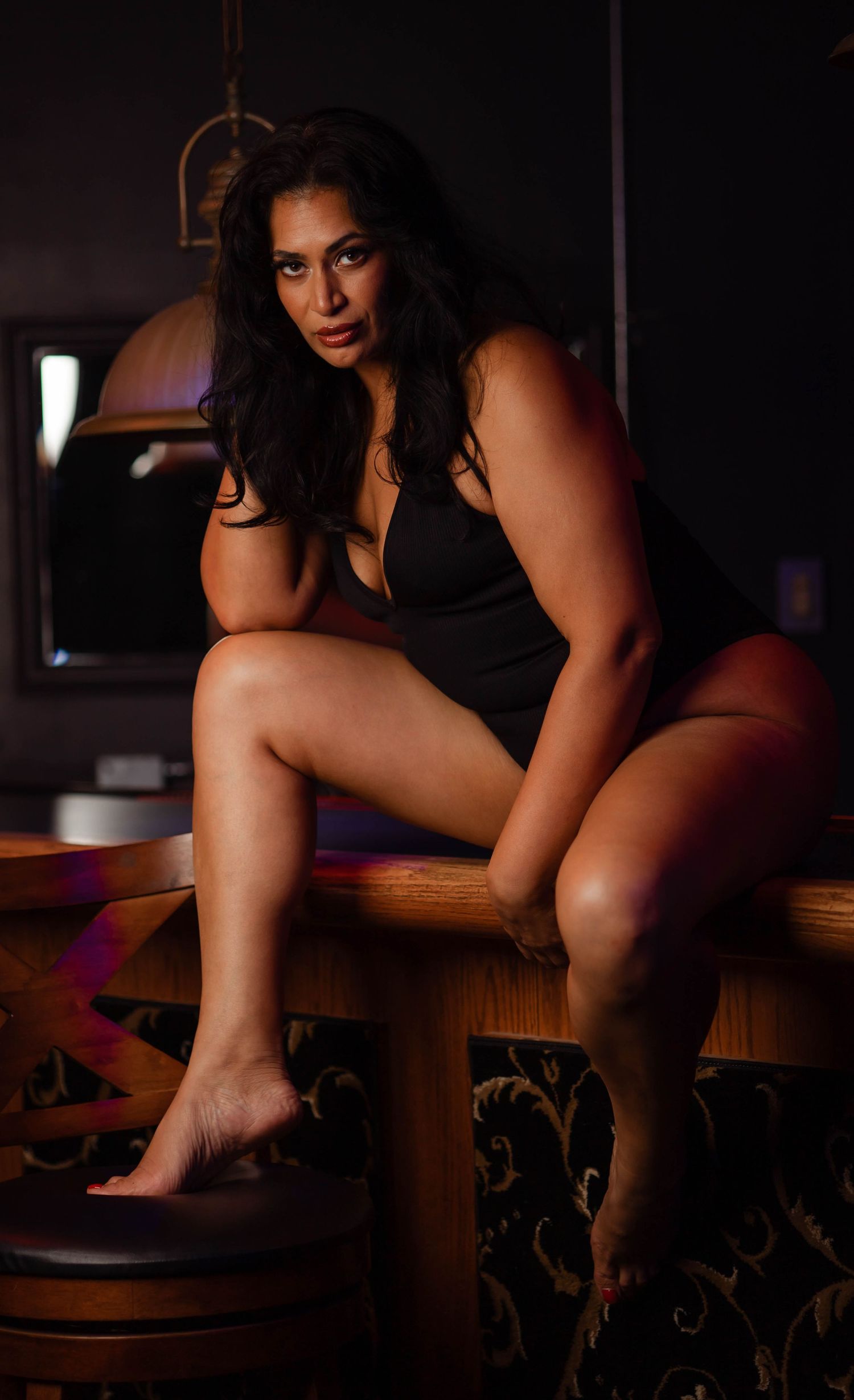 A woman in a black dress poses on a wooden table in a dimly lit room with moody lighting.