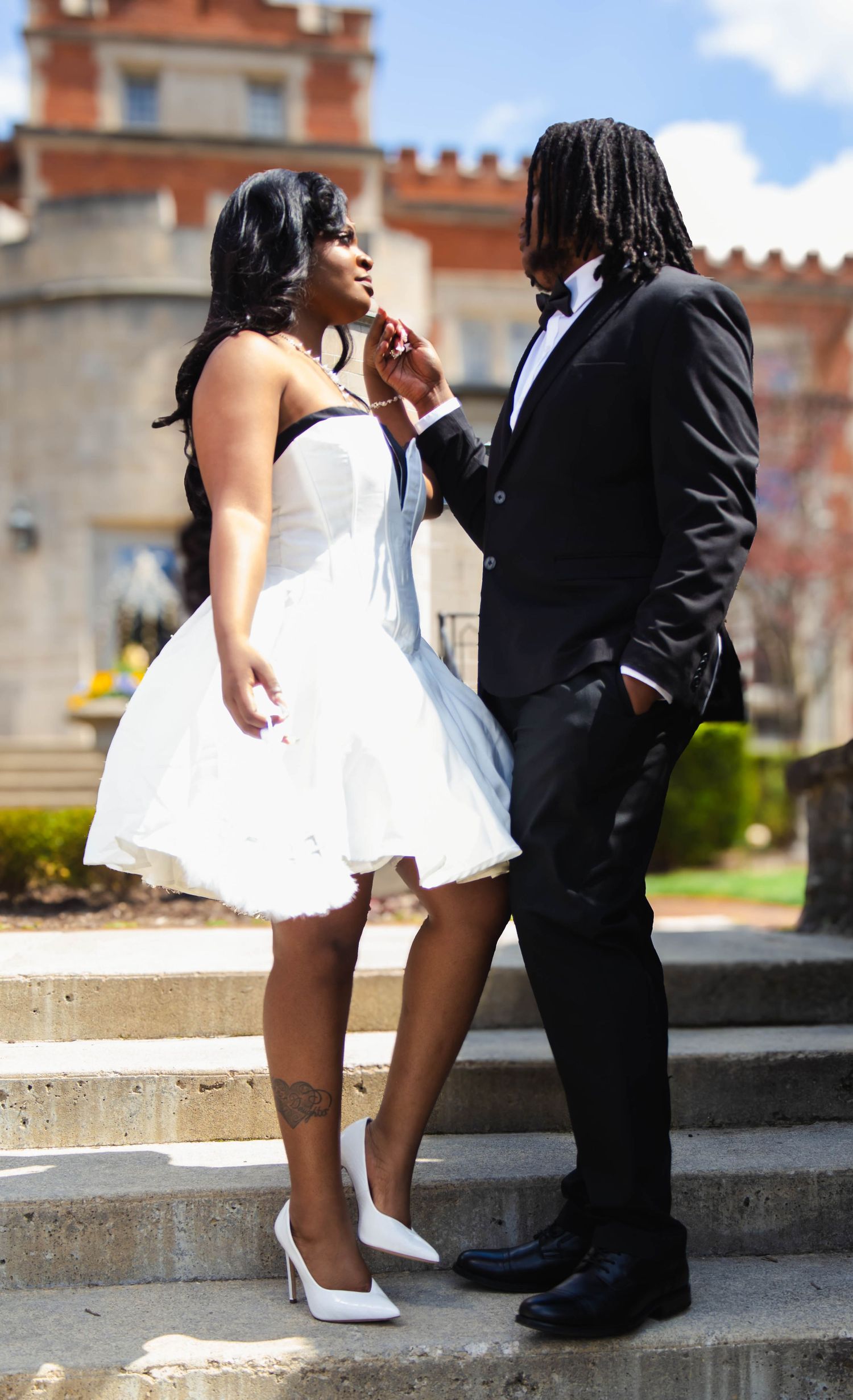 A couple dressed in formal attire share a romantic moment on stone steps outside a brick building on a sunny day.