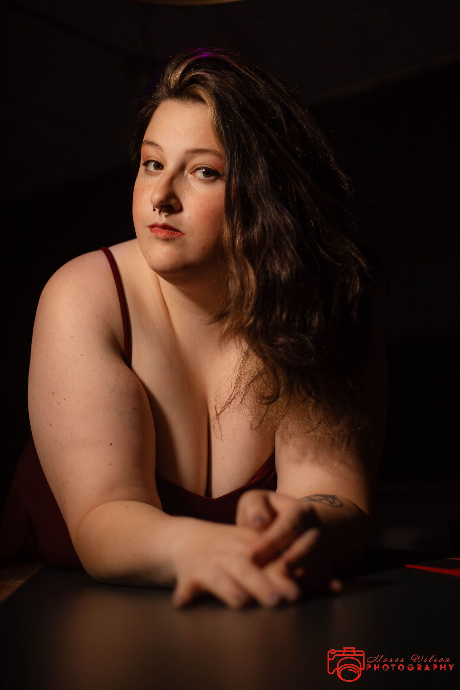 A young woman with dark wavy hair poses in a burgundy top, leaning on a surface in a dramatic studio portrait.