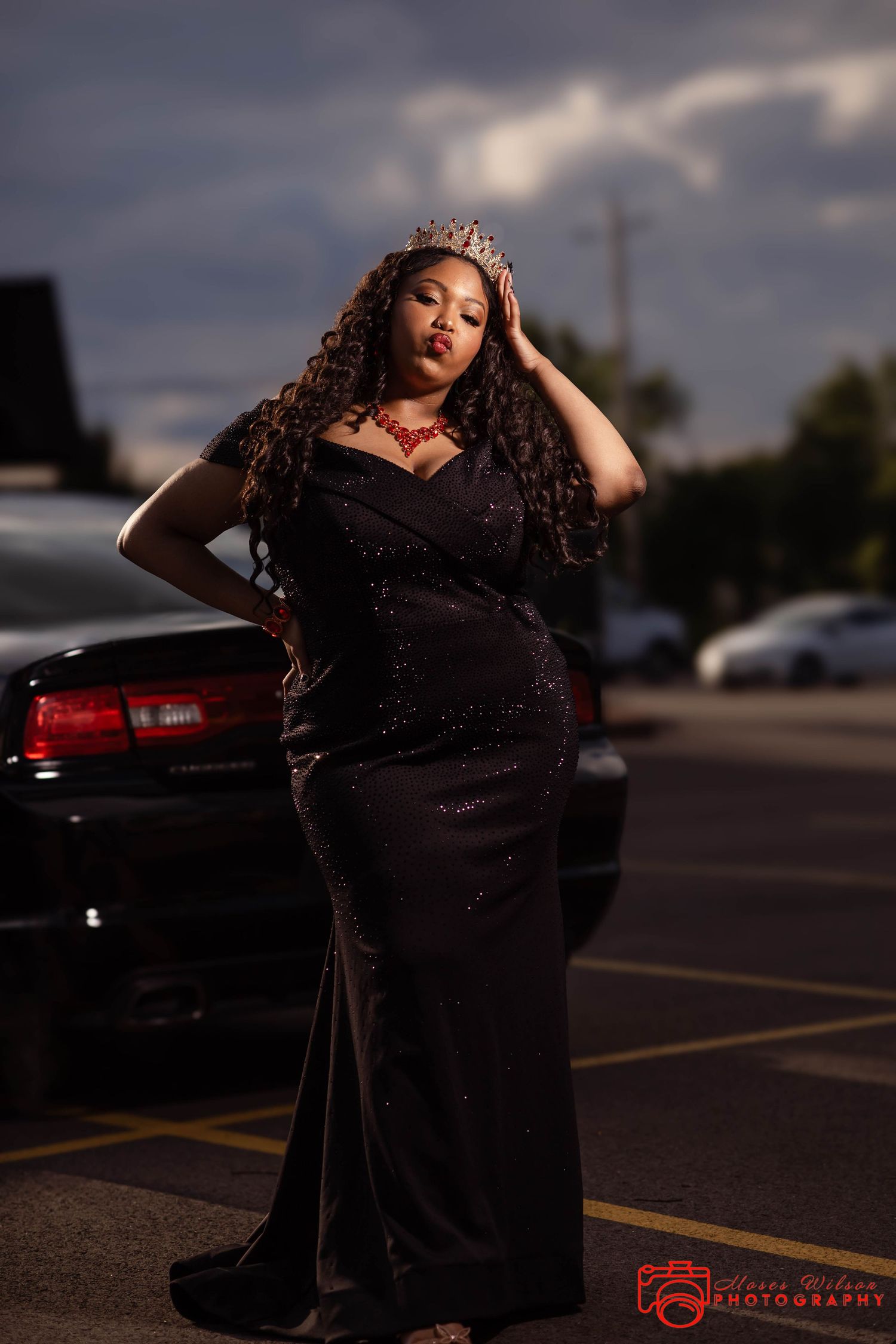 A plus-size woman in a black sequin gown and crown poses regally in a parking lot at dusk.