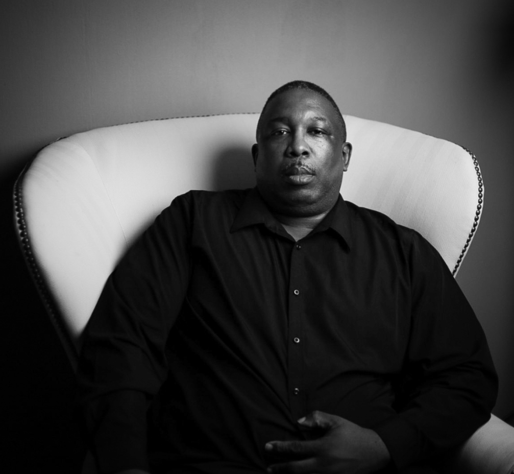 A moody black and white portrait of a person in dark clothing seated against a light chair.