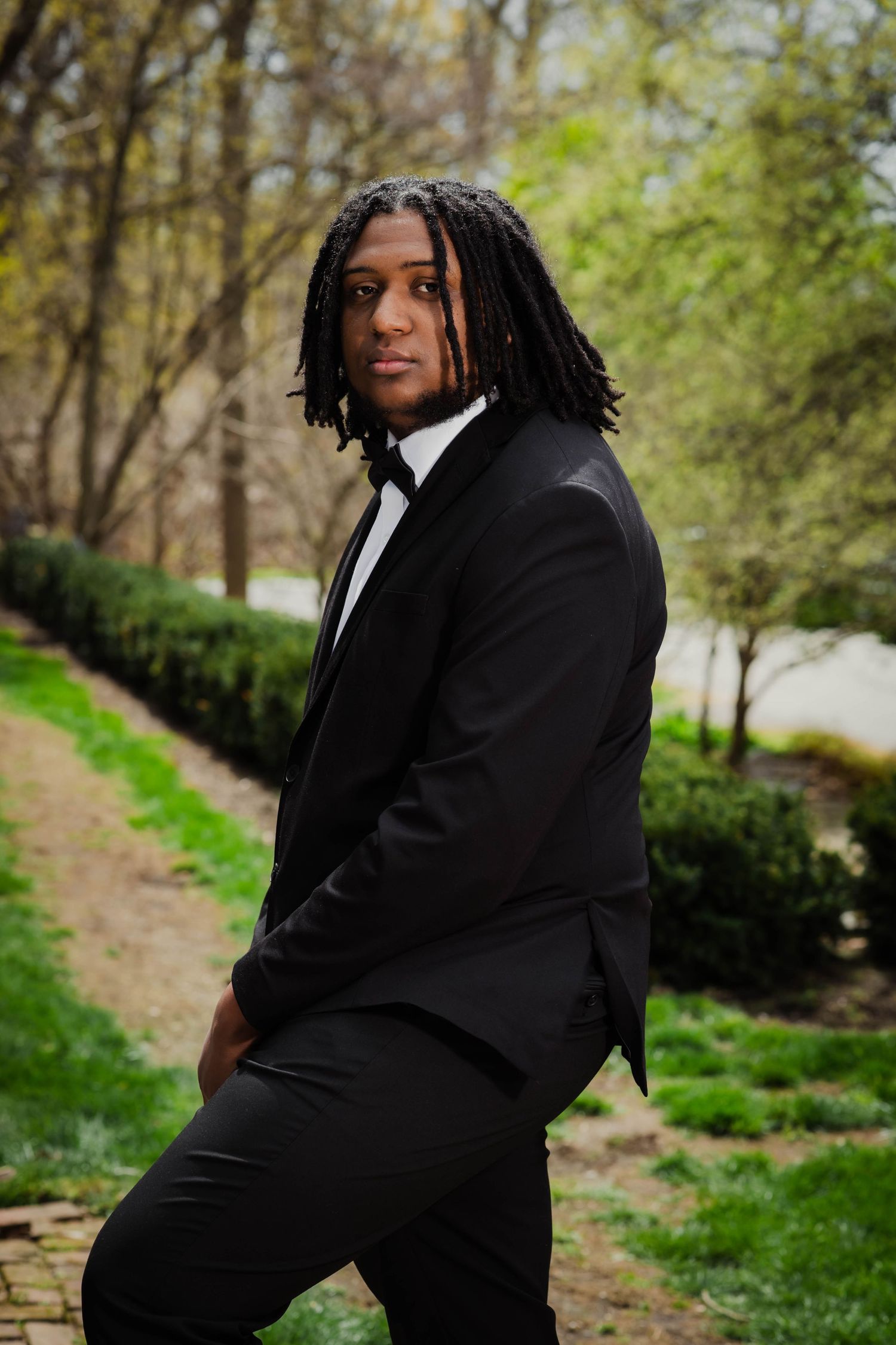 A formal portrait of someone in a black tuxedo posing on garden path.