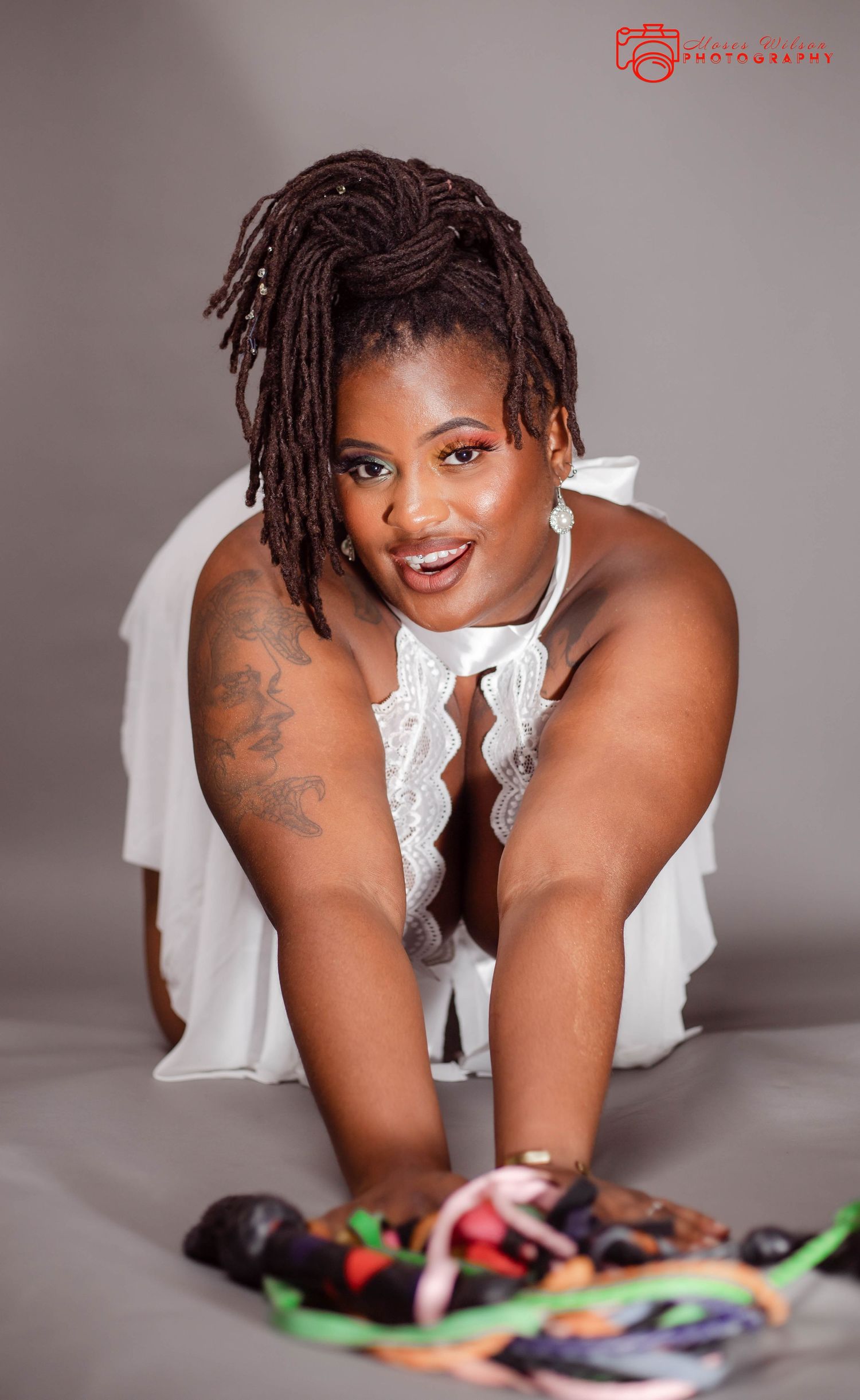 A person in a white lace dress leaning forward with dreadlocks hairstyle and tattoos, smiling at the camera.