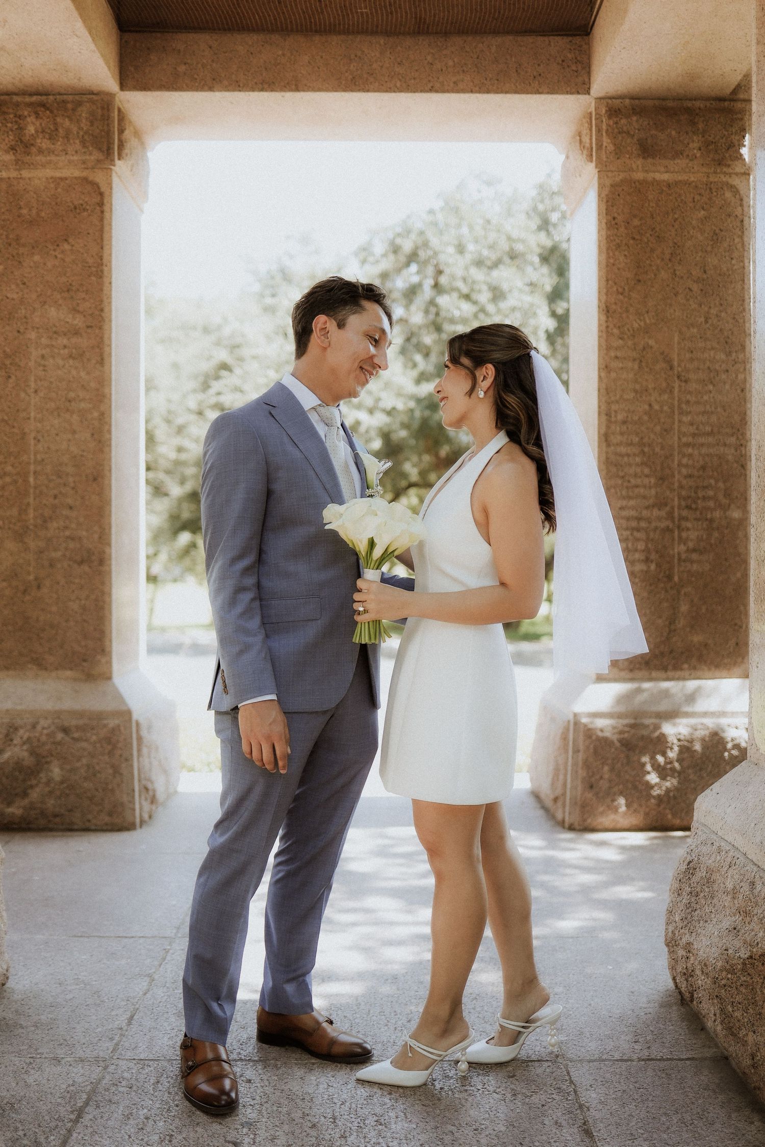 A couple in wedding attire shares an intimate moment under stone columns in a romantic outdoor setting. texas capitol