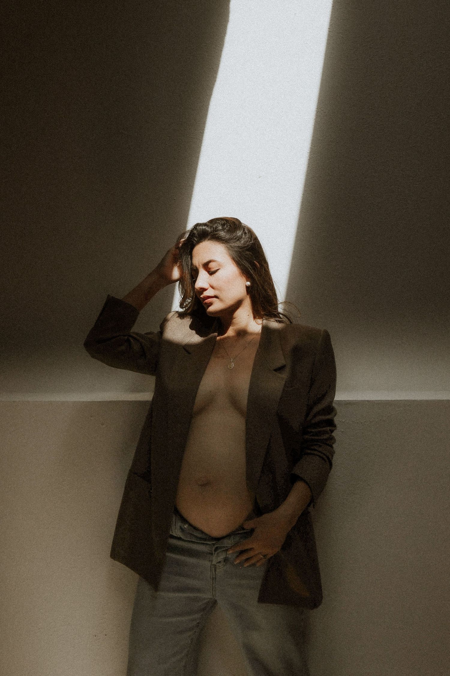 A figure in a dark blazer and neutral top poses dramatically against a wall with stark shadows and lighting. maternity, mom to be