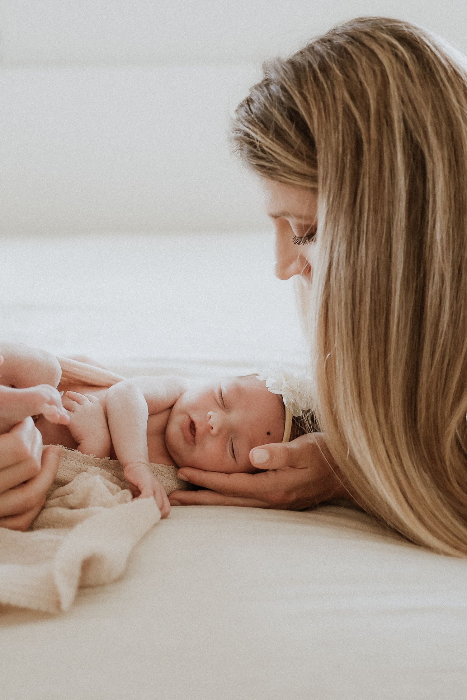 A tender moment between parent and newborn baby lying together on white bedding in soft natural light.