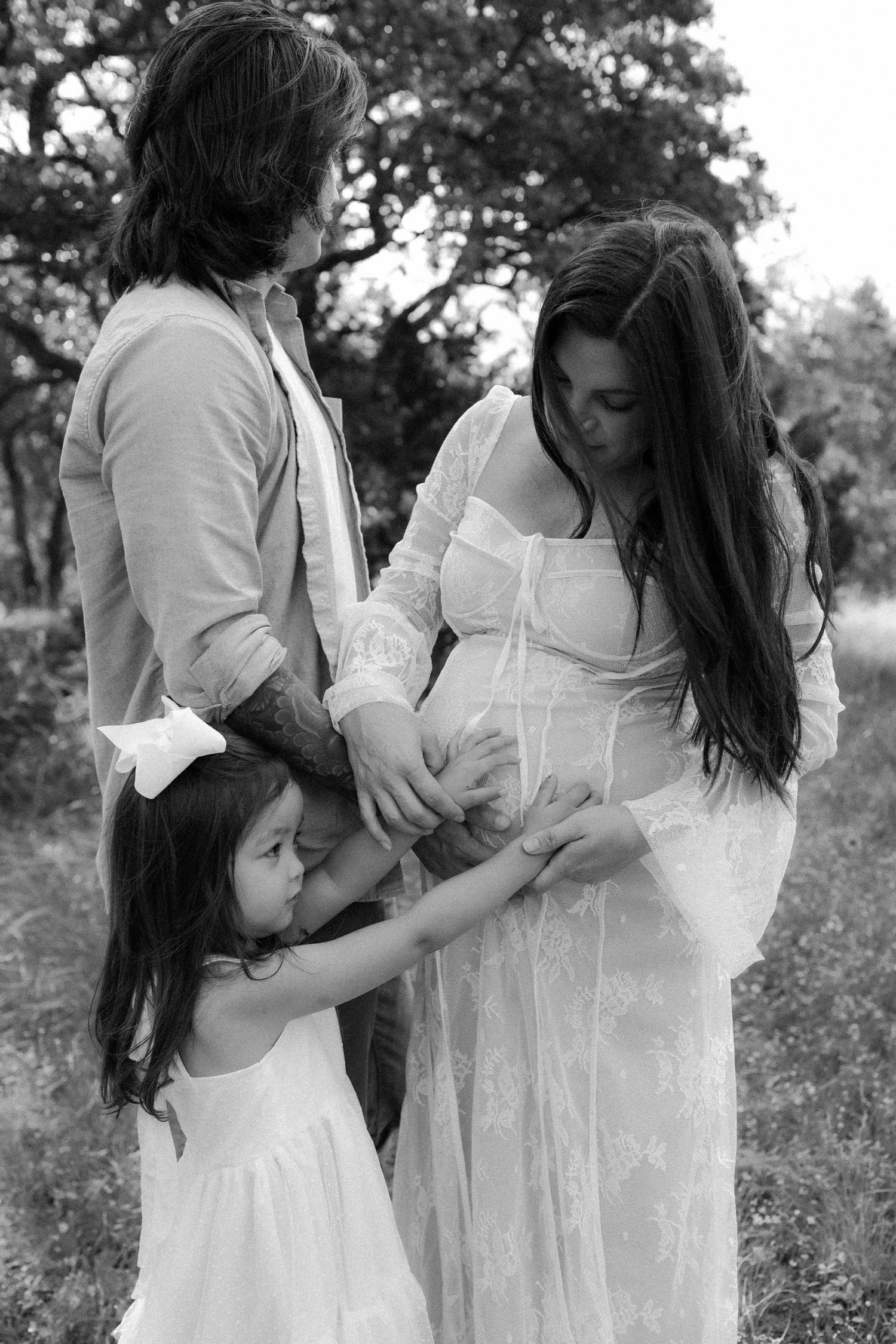 A family in white clothing stands together outdoors in a tender black and white maternity photo. texas hill country