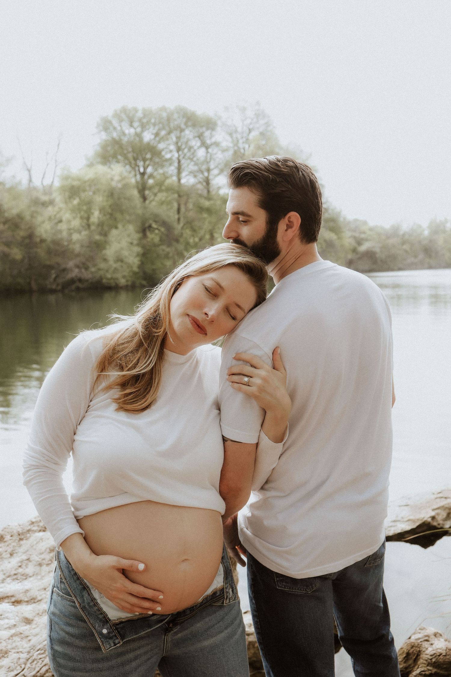 Intimate maternity photo session by a lake showing couple in white shirts sharing tender moments together.