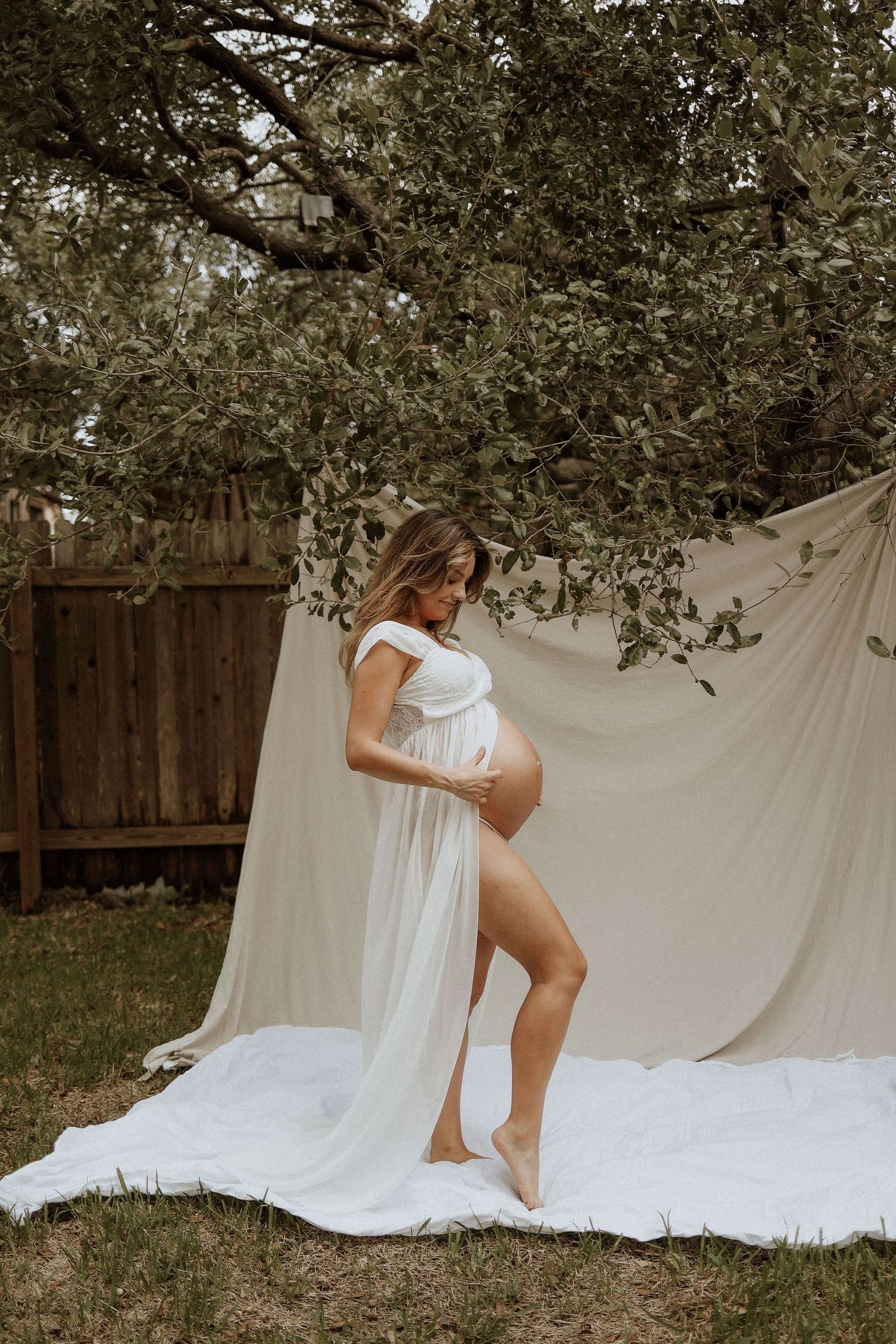Artistic maternity photo outdoors with white fabric backdrop and flowing dress under a tree in natural lighting. backyard, photoshoot