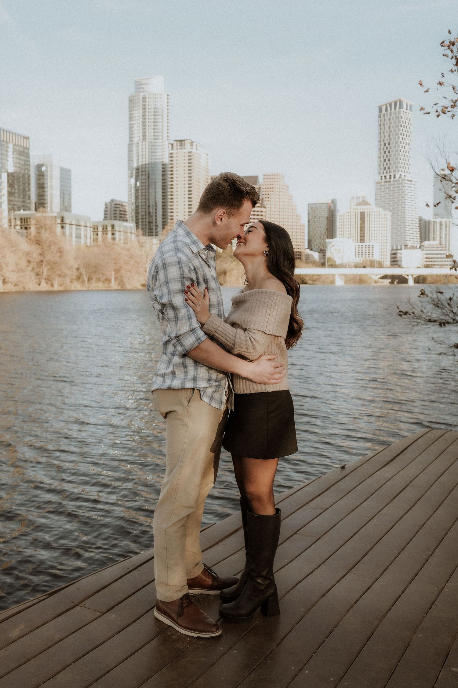 A romantic couple shares a tender kiss on a wooden pier overlooking a city skyline and waterfront at sunset. auditorium shores, austin, tx