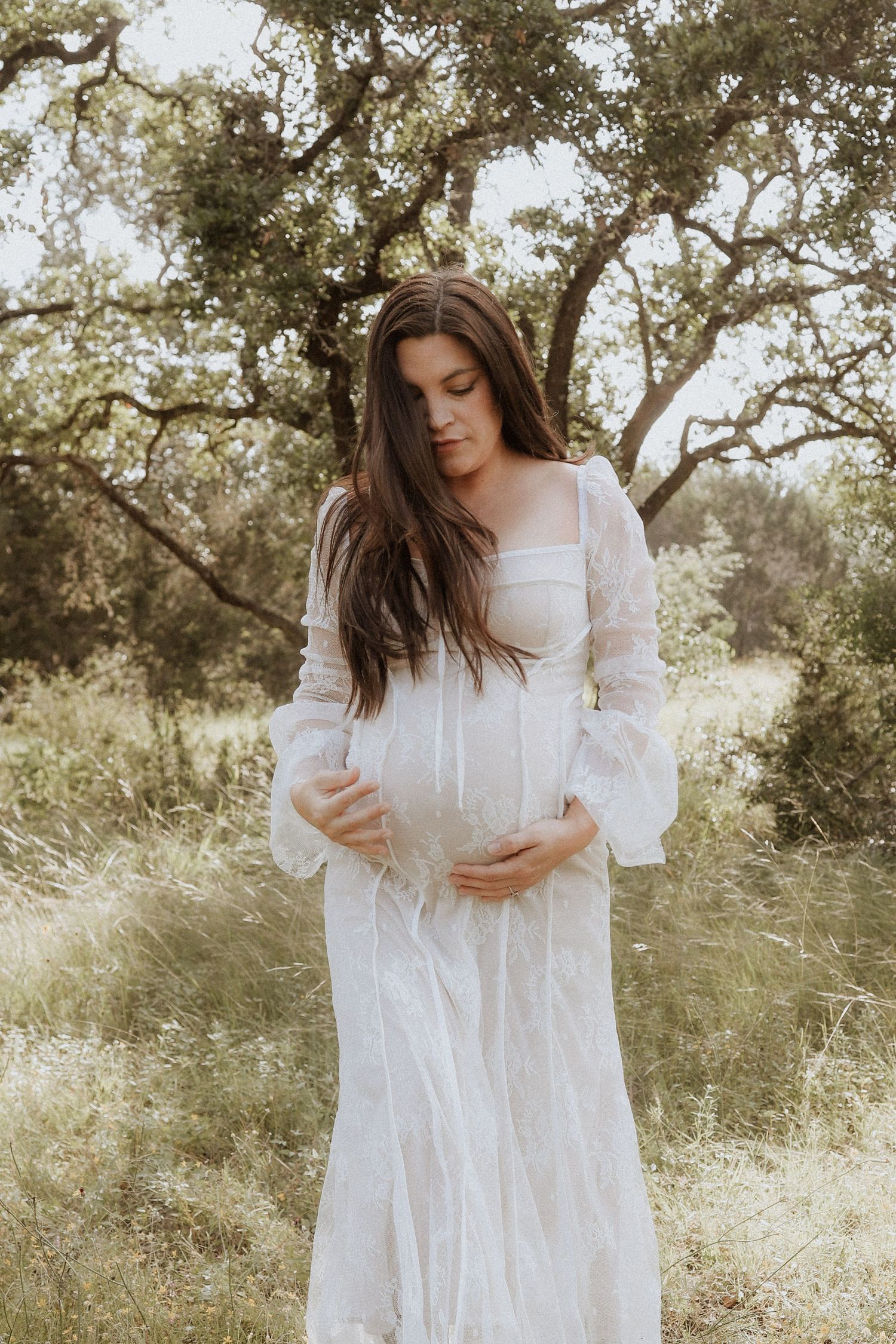 A person in a flowing white lace dress stands among sunlit trees in a dreamy outdoor setting. maternity photoshoot texas hill country