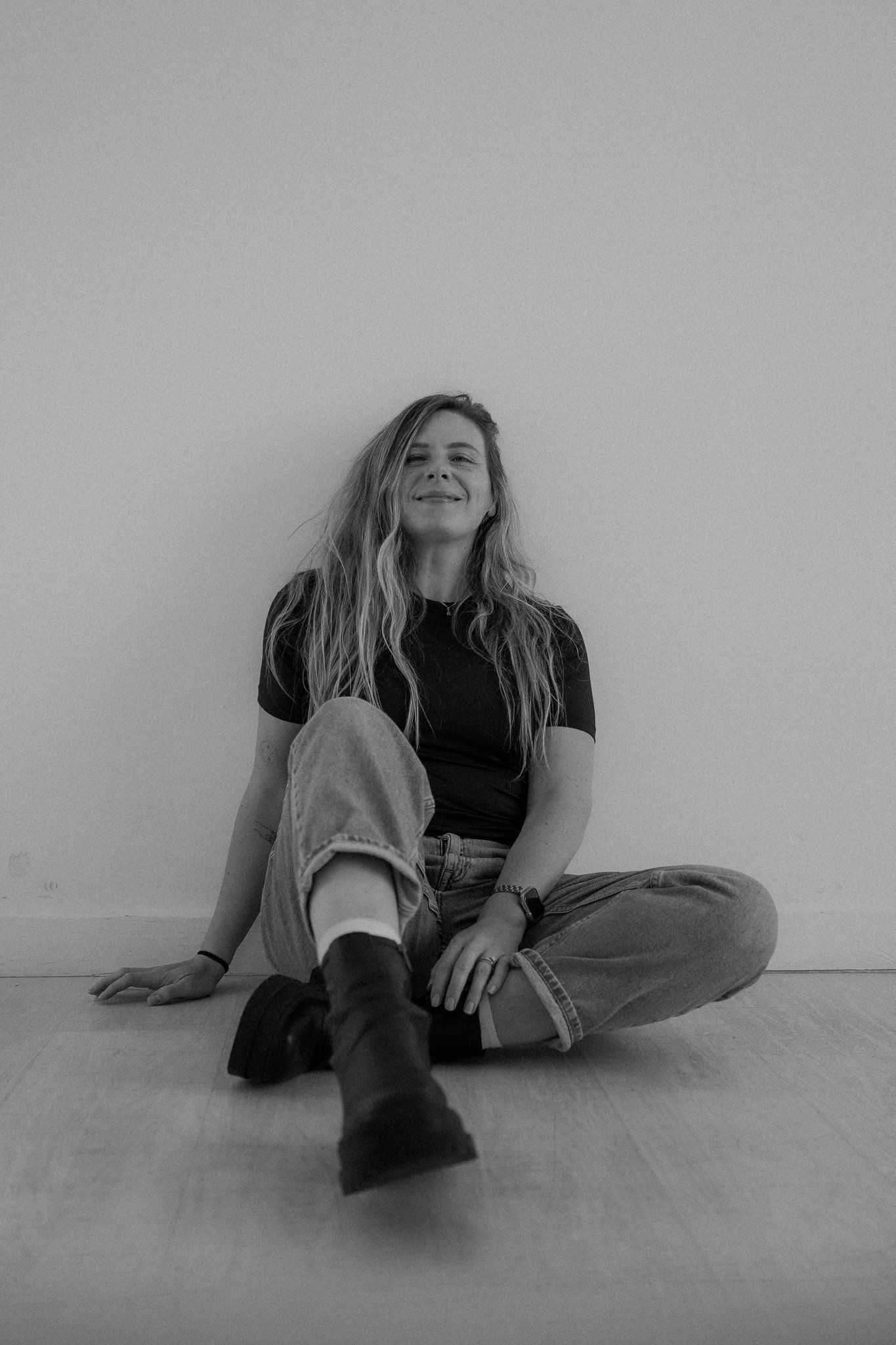 Black and white portrait of someone sitting on floor against wall wearing casual clothes and smiling upward.