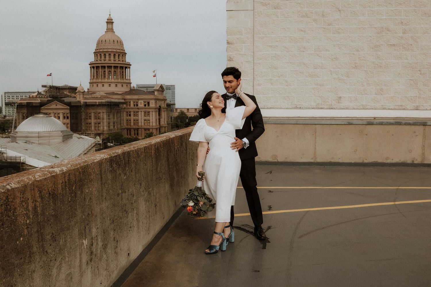 A couple in formal attire poses on a rooftop with the Texas State Capitol building visible in the background, downtown austin, wedding