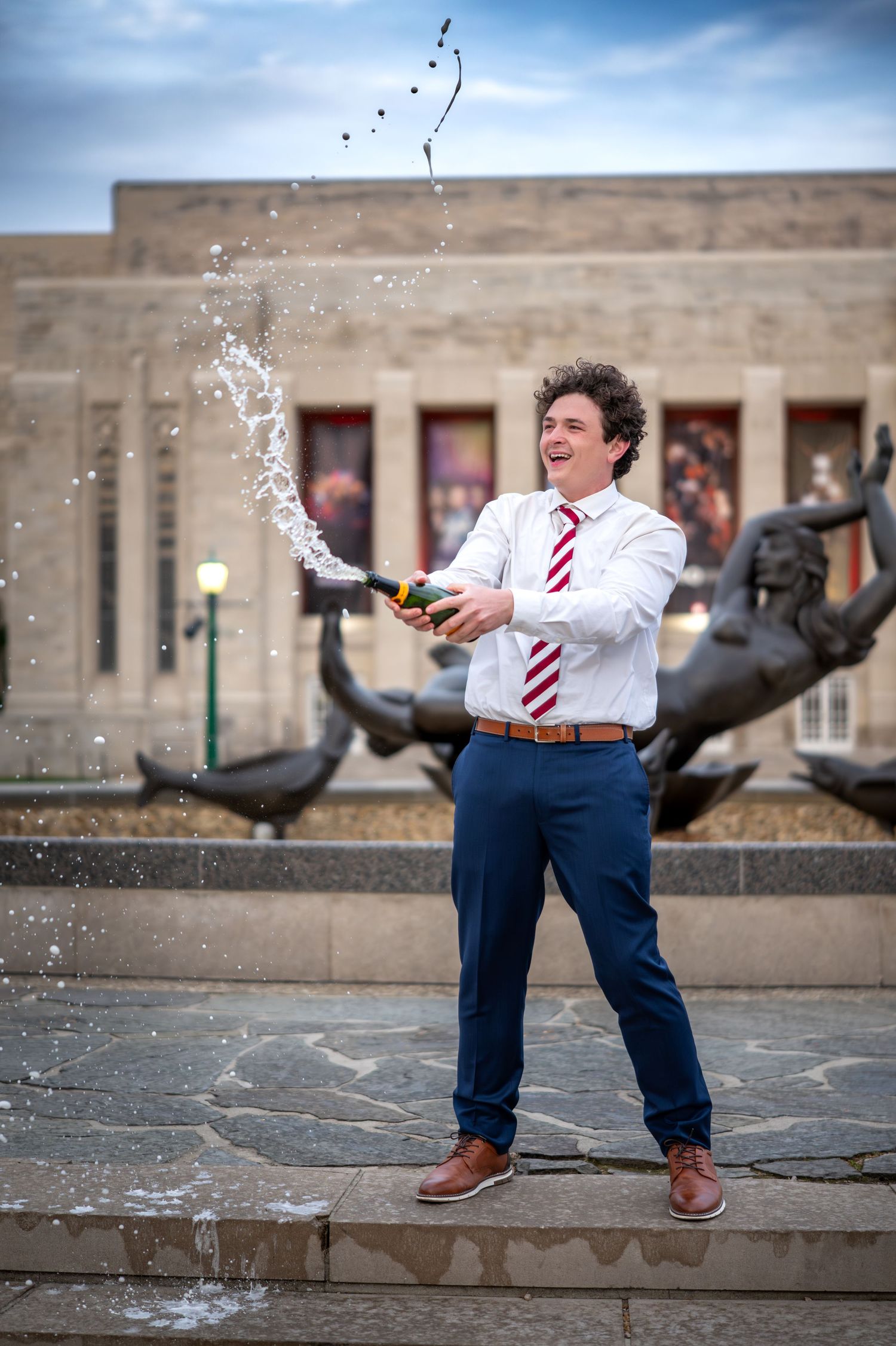 Indiana University graduate celebrating by shaking champagne at Showalter Fountain in Bloomington, wearing cap and gown.
