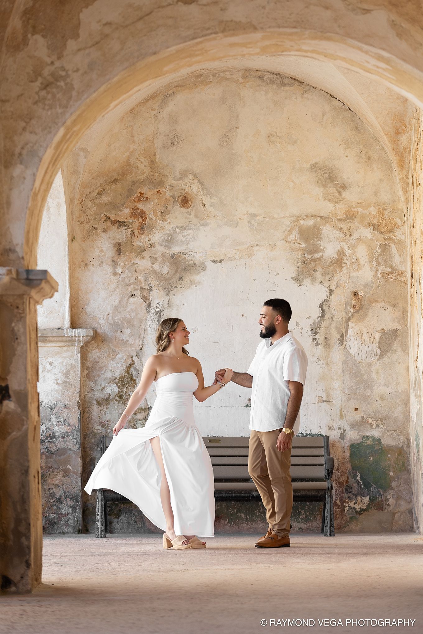 Couple dancing under stone arch at historic venue, woman in white gown, man in khaki, by Raymond Vega Photography. Castillo de San Cristóbal