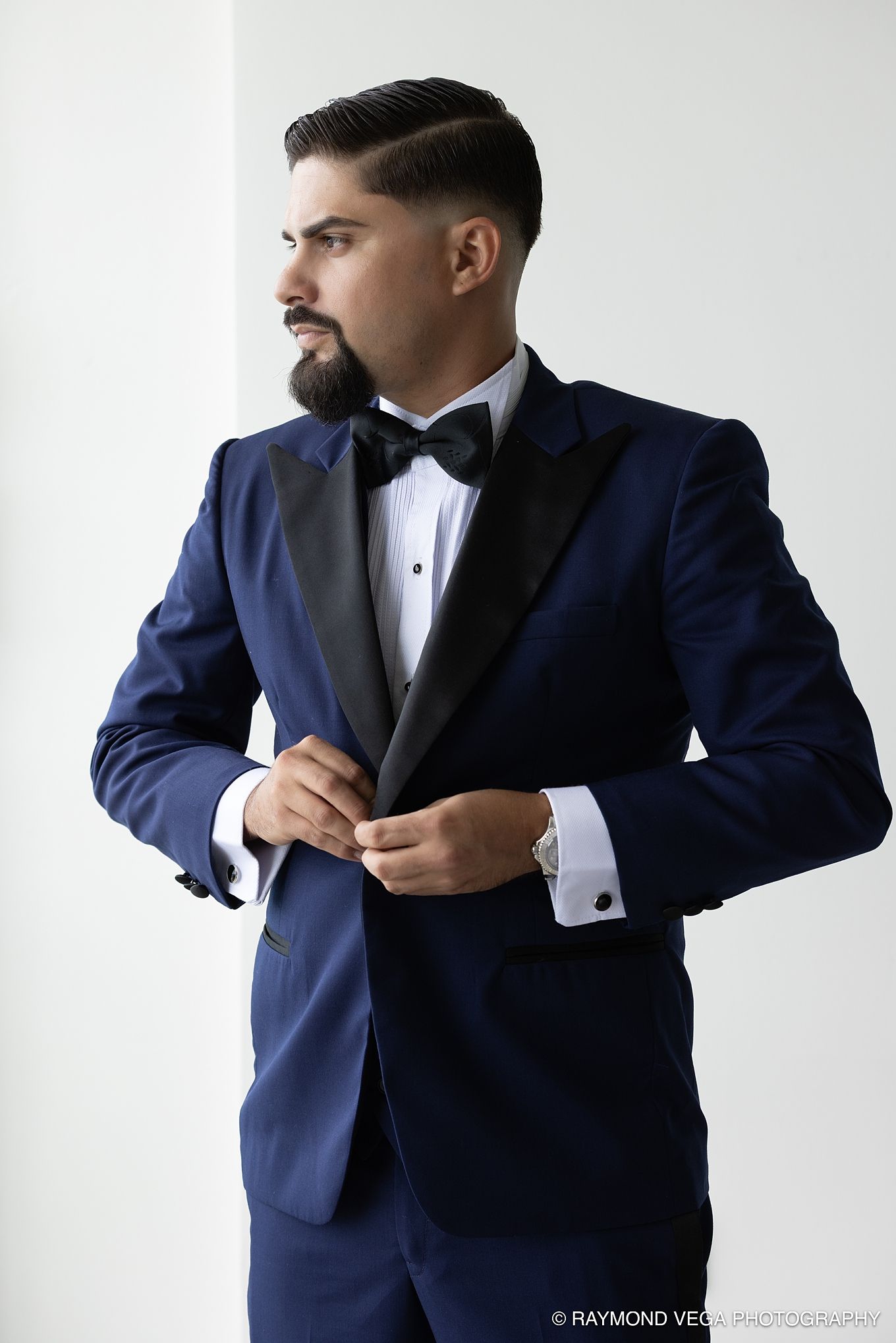A man in a navy blue tuxedo with black lapels and bow tie buttoning his jacket in a bright white studio. Hotel la Concha Wedding