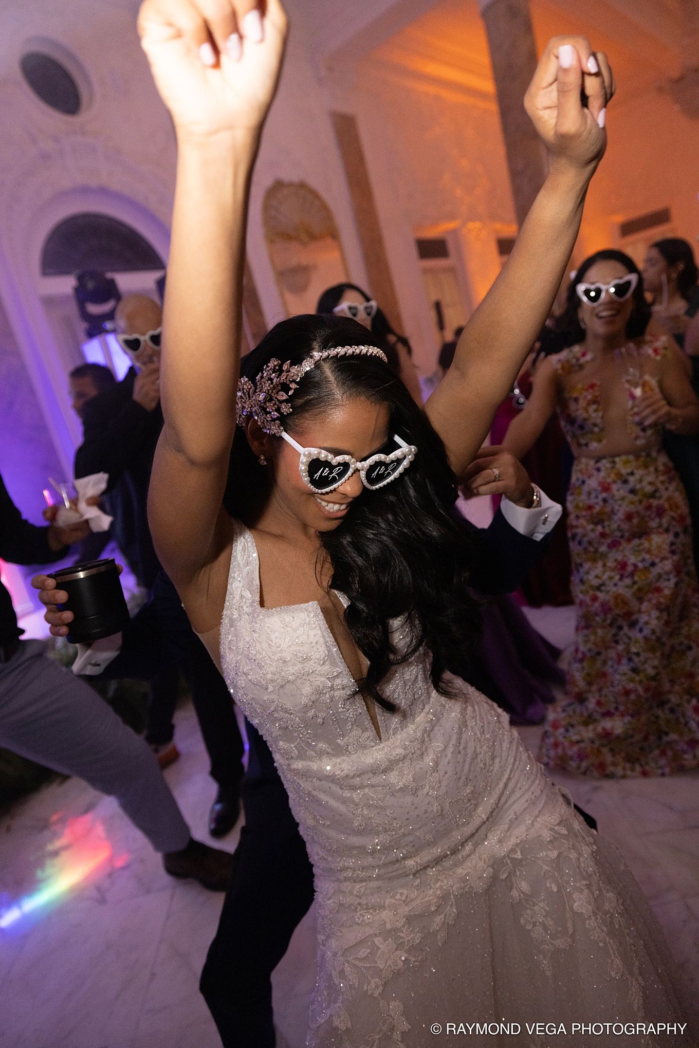 Bride in white lace gown and heart-shaped sunglasses dances joyfully at wedding reception with arms raised.Antiguo Casino Puerto Rico weddin